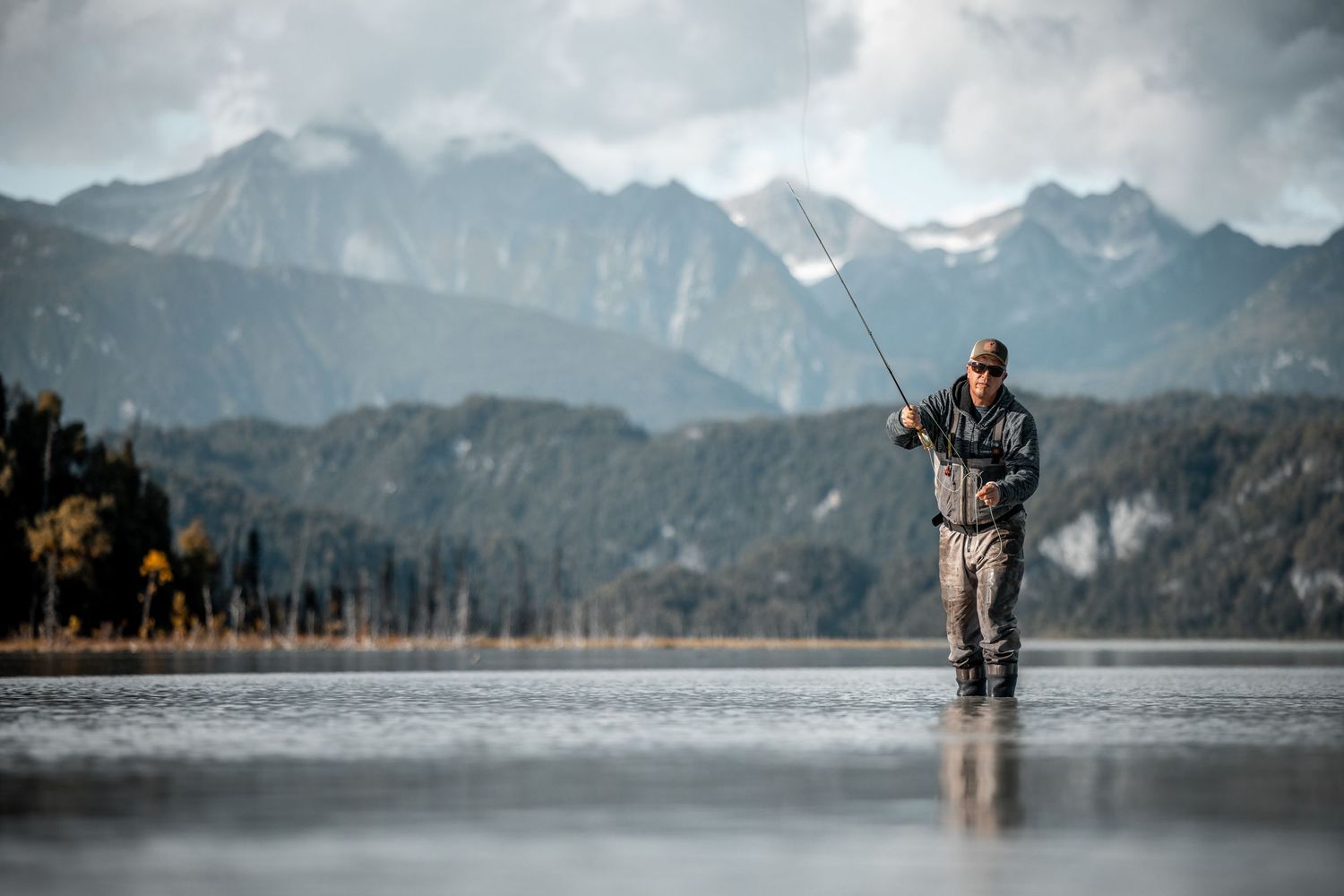 Fly fisherman standing in mountain lake with snow-capped peaks in background.