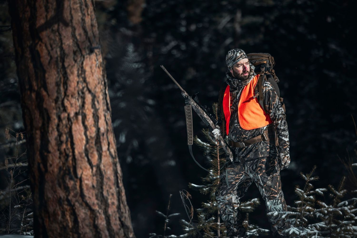A hunter in orange safety vest and camouflage stands in a dark forest with rifle.