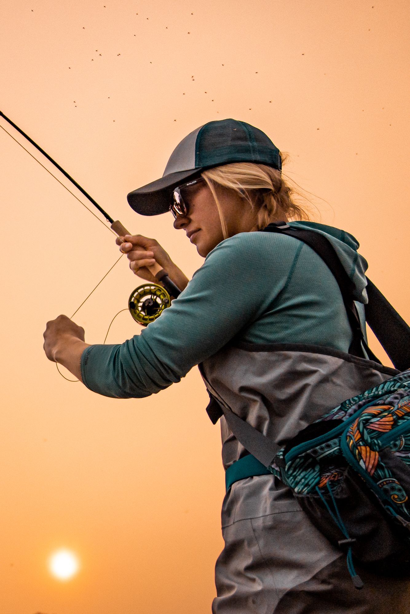 Fly fisher casting a line at sunset with fishing gear and backpack silhouetted against an orange sky.