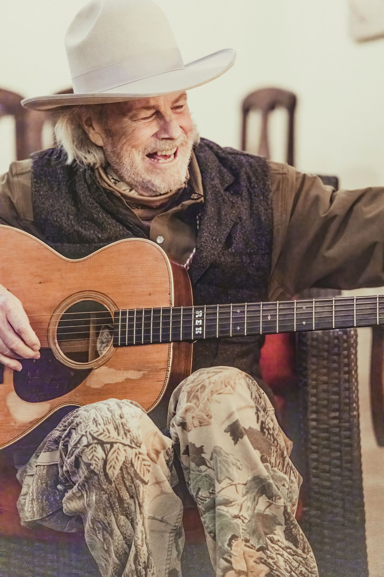 Musician wearing cowboy hat and camo pants plays acoustic guitar indoors.