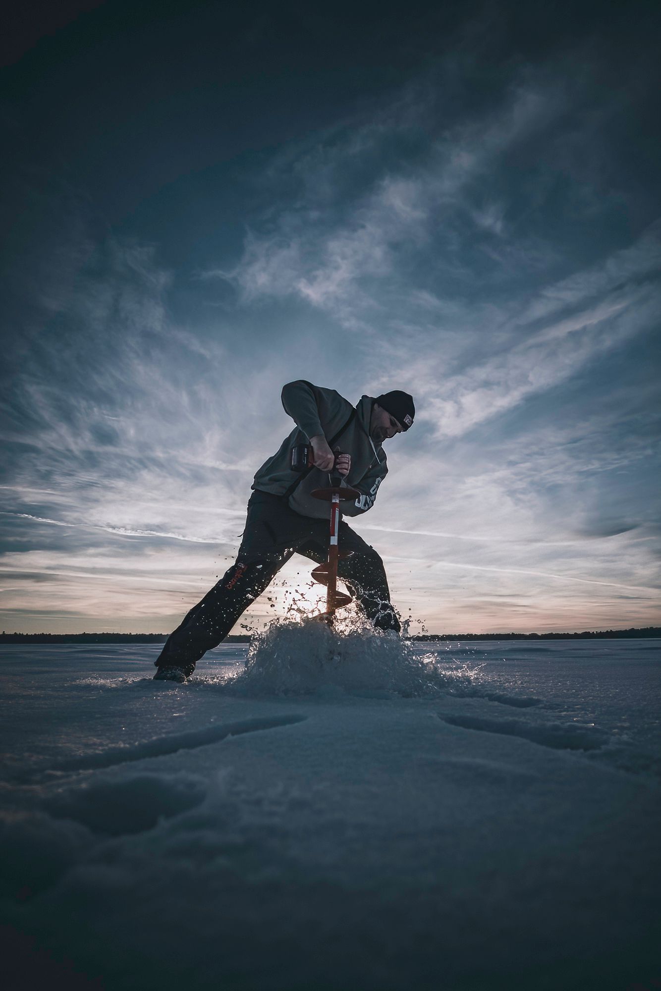 Dynamic shot of person drilling ice hole with auger against dramatic winter sky.