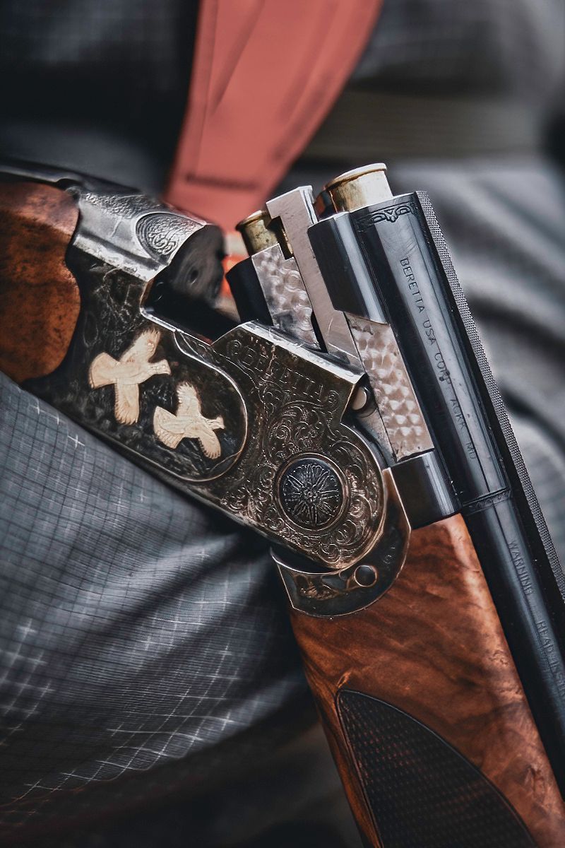 Close-up details of an ornately engraved shotgun with wooden stock in dramatic lighting.
