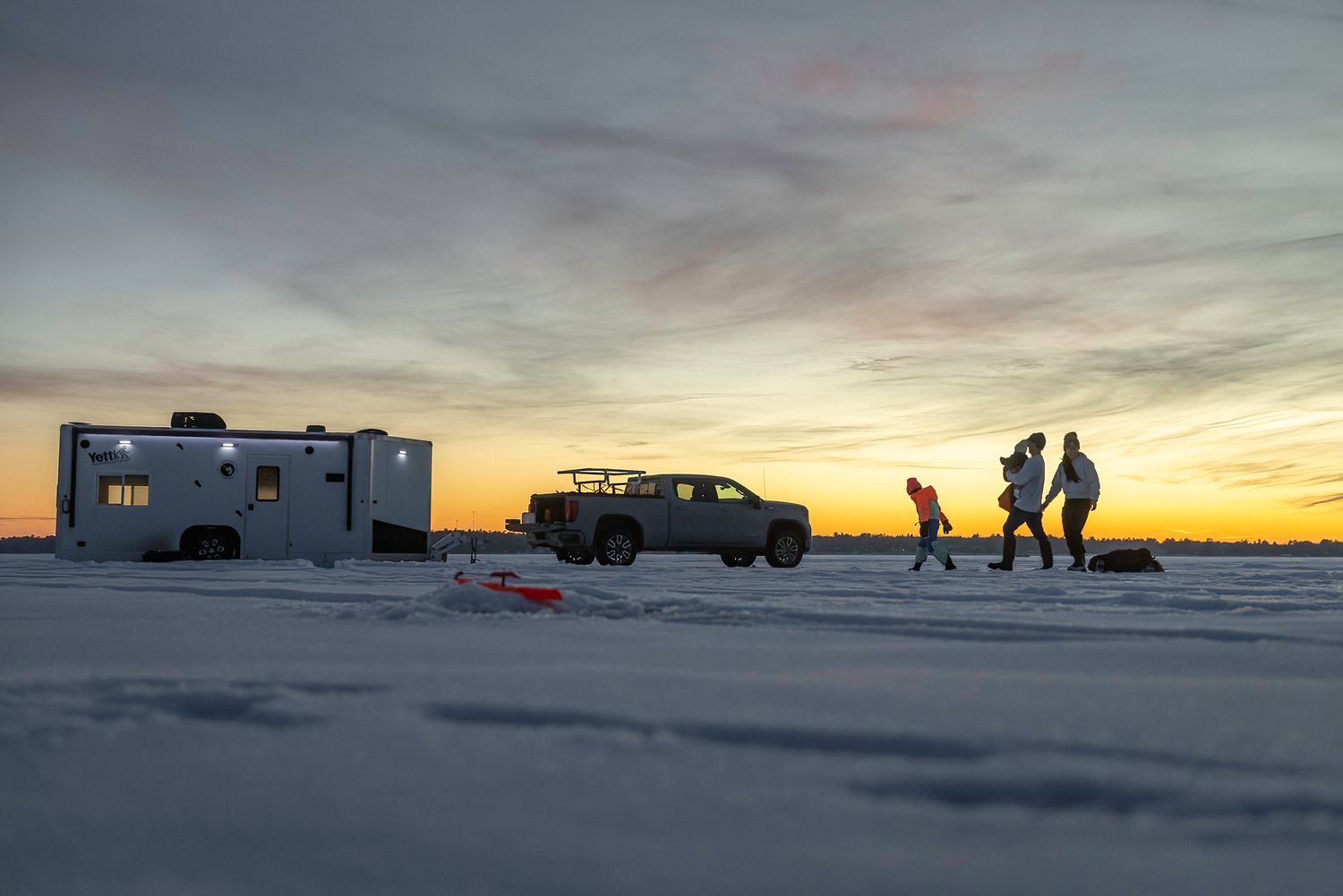Silhouettes of people and RV against vibrant winter sunset on a snow-covered landscape.