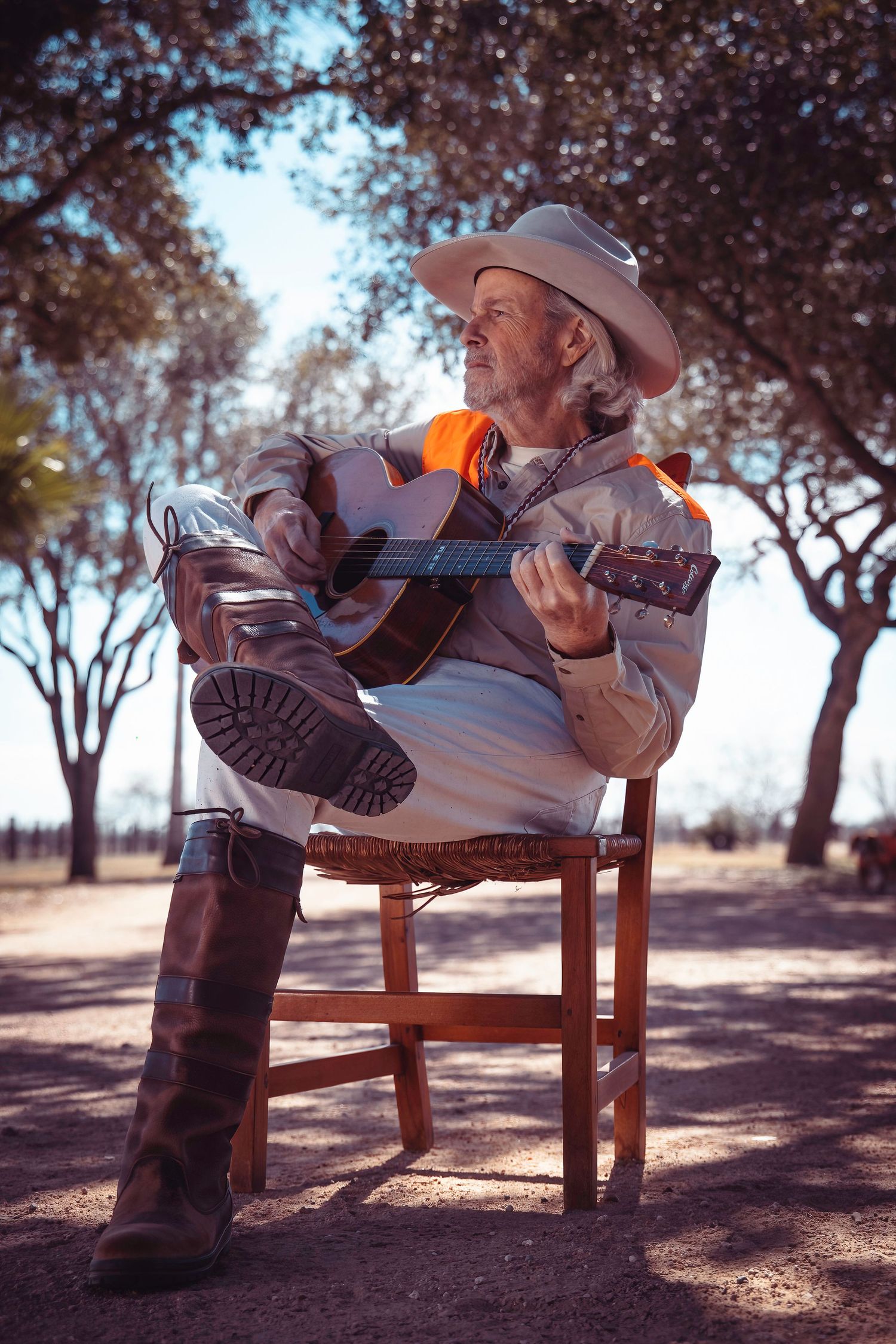 Person relaxing on wooden chair playing guitar with boots propped up in outdoor setting.