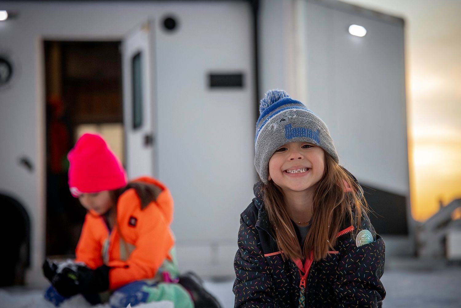 Winter scene with people in warm clothing sitting outside an RV in snowy conditions.
