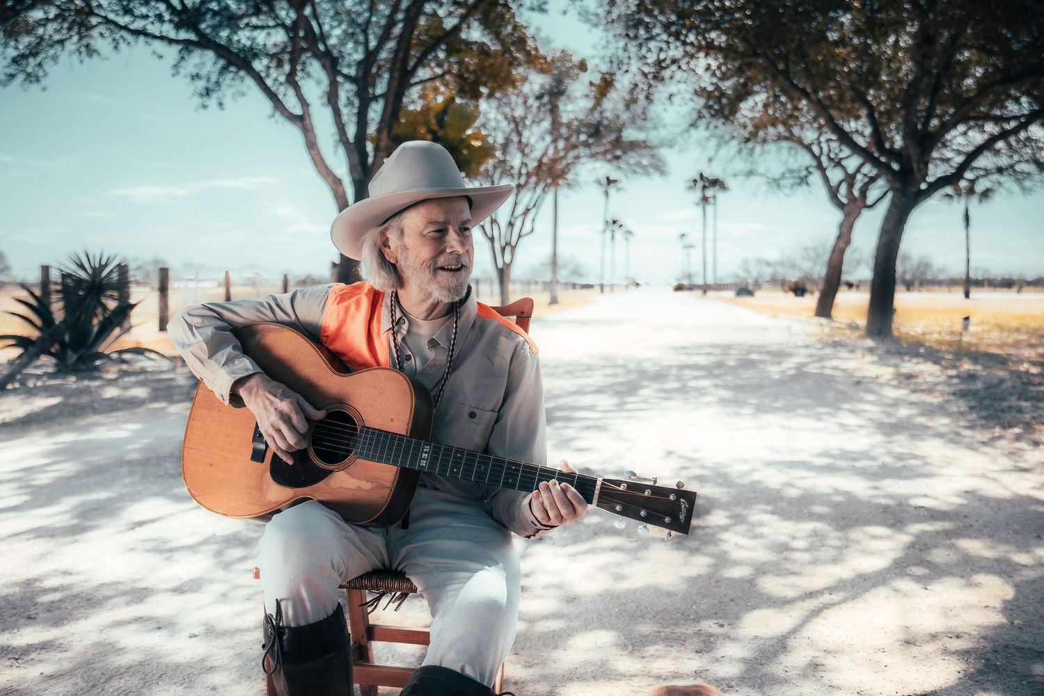 Musician in orange shirt and cowboy hat plays acoustic guitar outdoors under shaded trees.