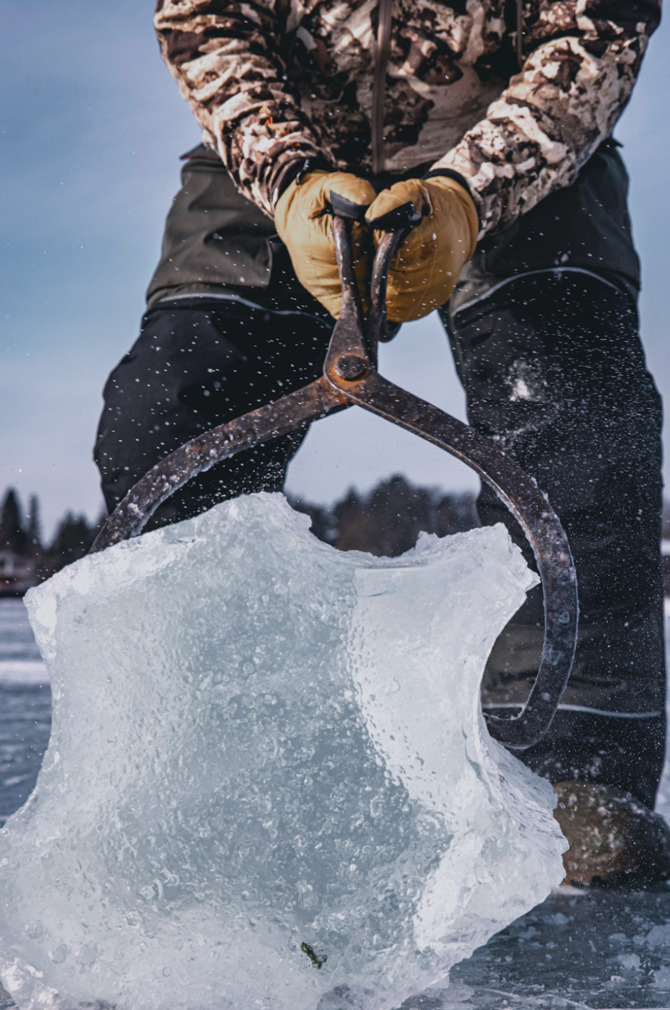 Person using ice tongs to grip large chunk of clear ice on frozen lake.