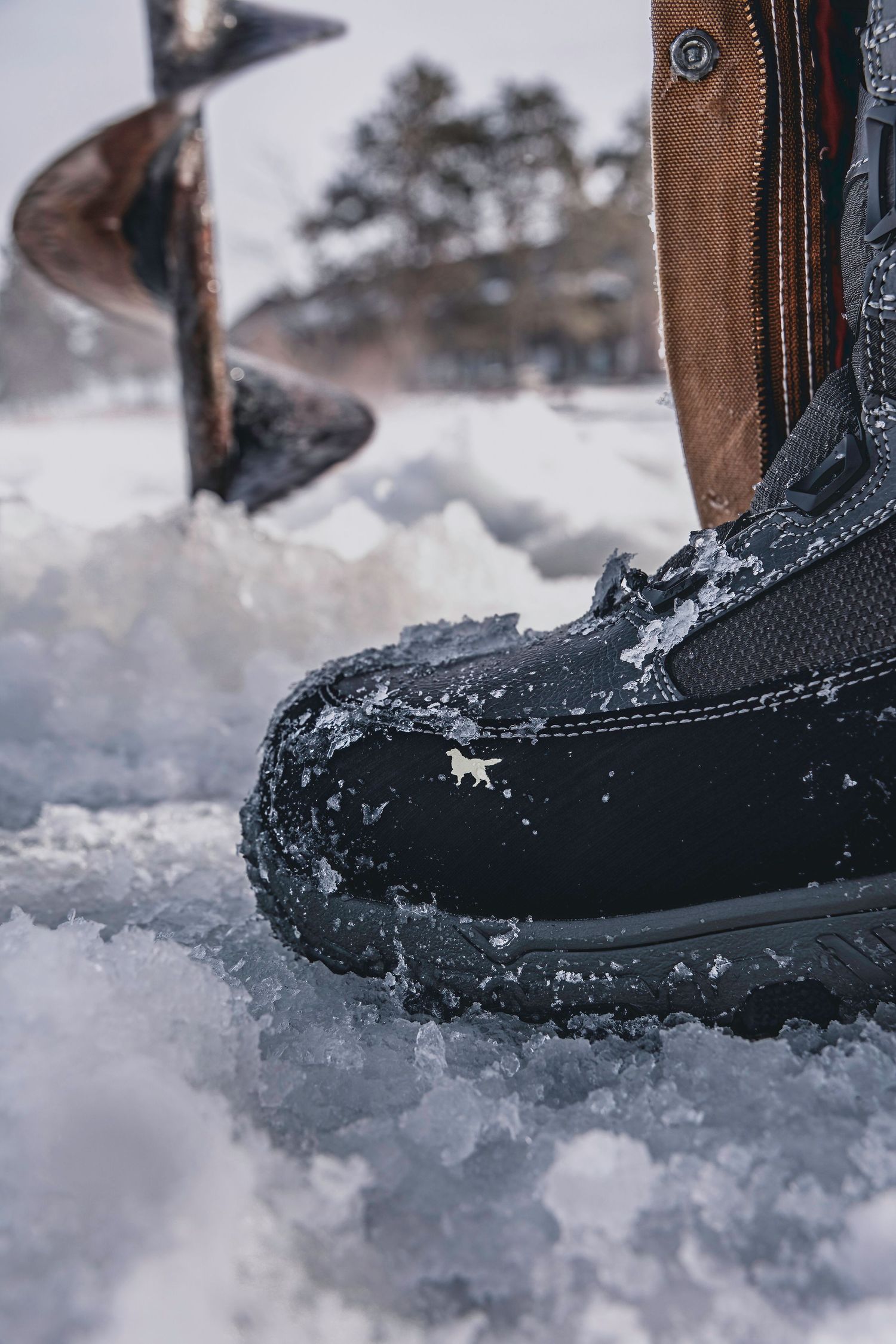 Snow-covered winter boot next to ice fishing hole on frozen lake surface.