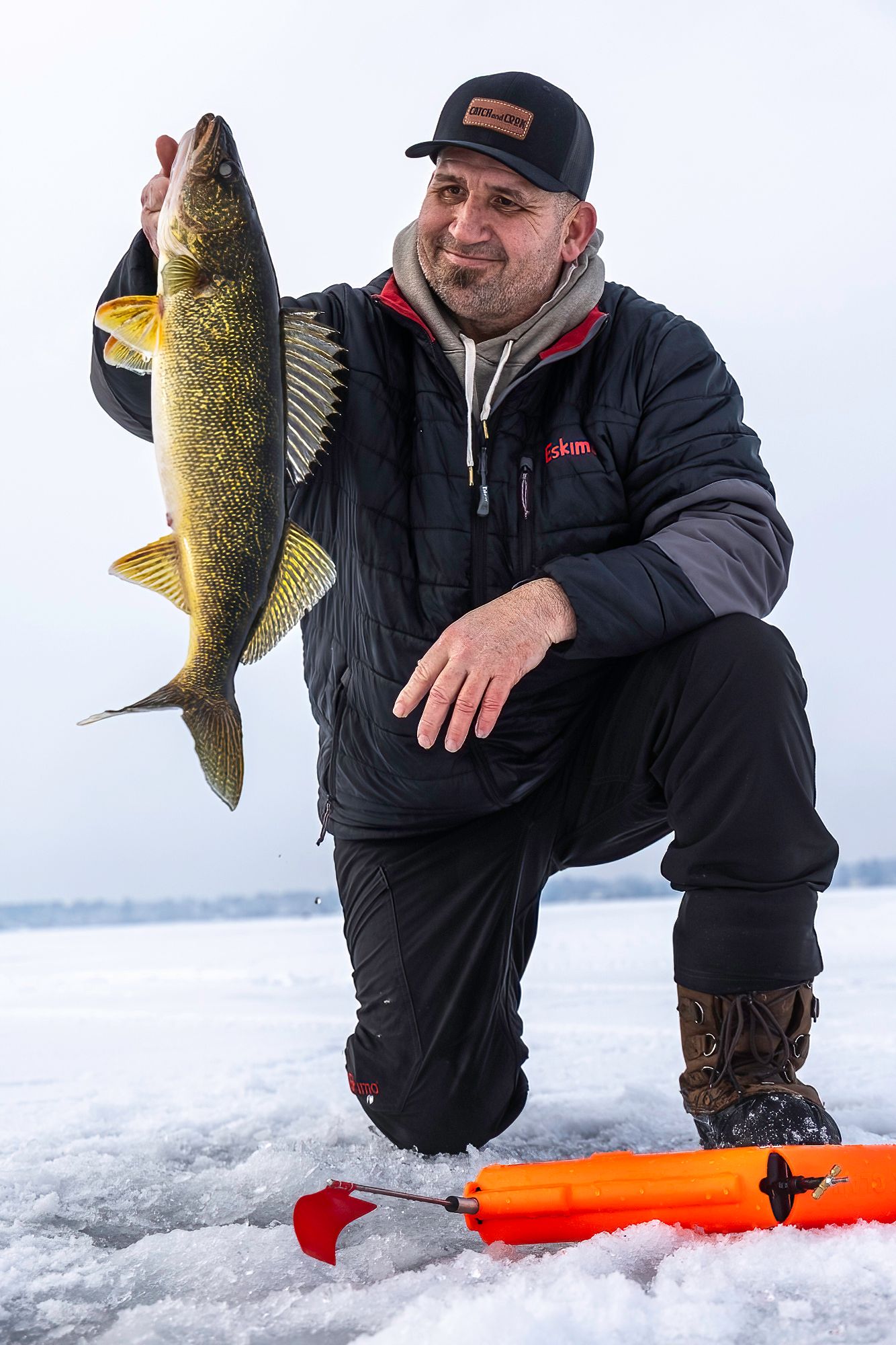 Angler in black winter gear displays a freshly caught walleye through an ice fishing hole.