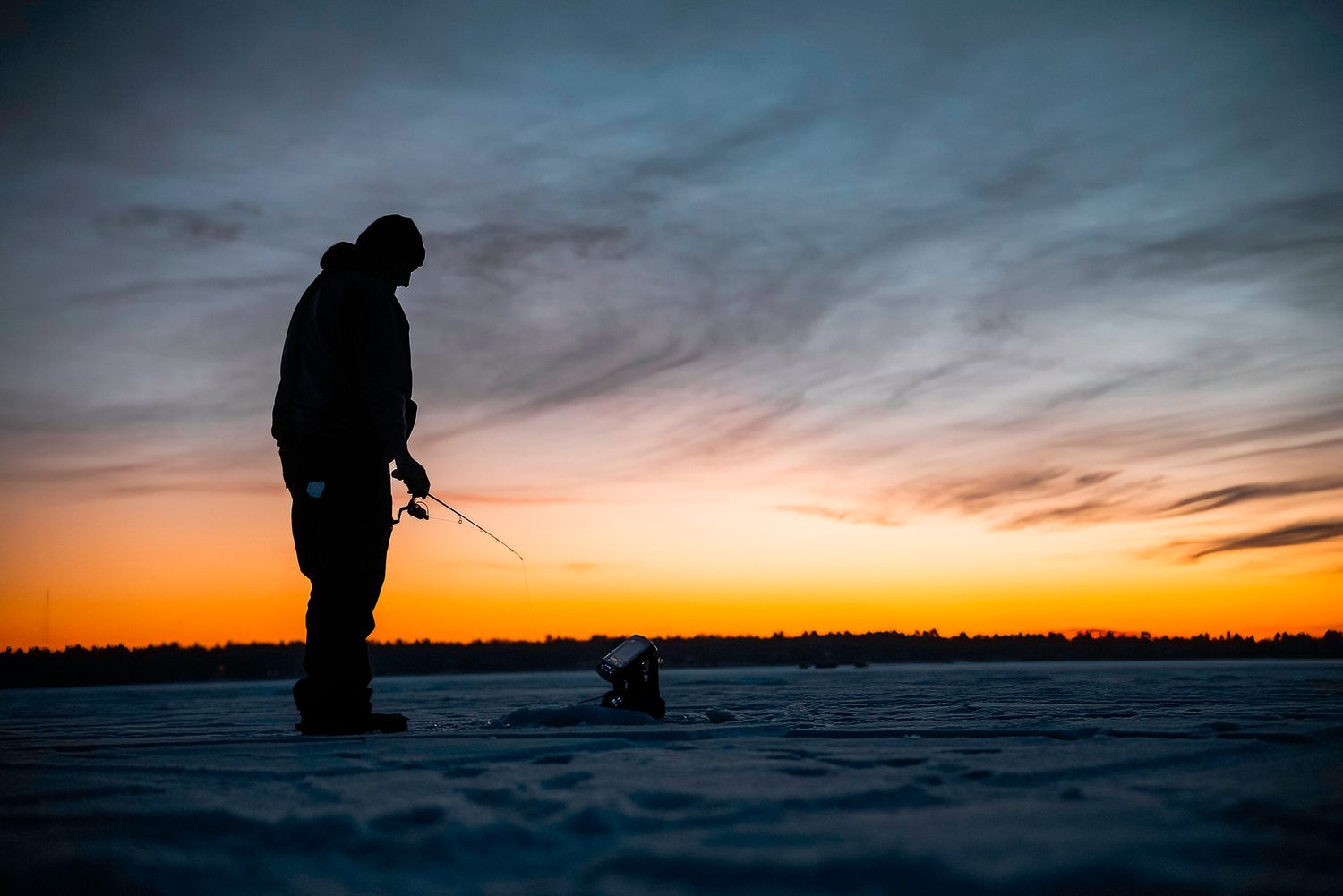 Silhouette of ice fisherman standing on frozen lake during dramatic orange sunset.