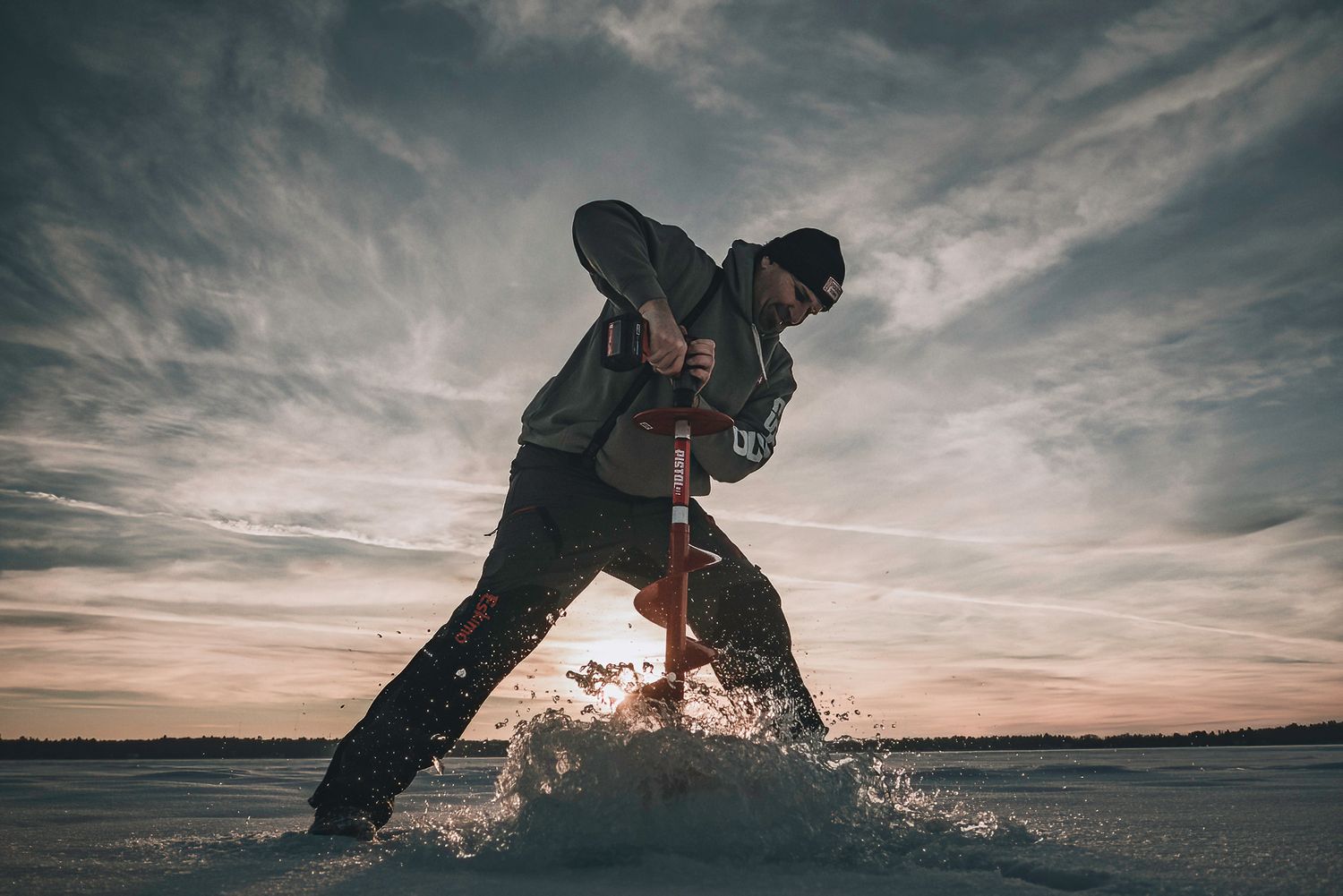 Ice fisherman drilling a hole in frozen lake at sunset with dramatic clouds and splashing ice.