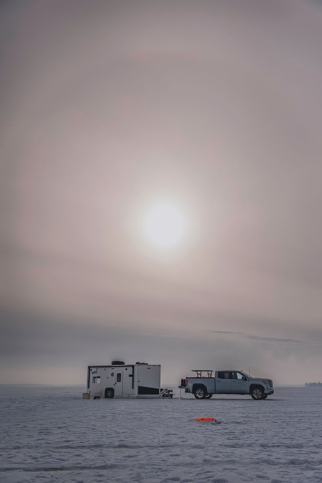 Ice fishing shelters and vehicles dot the frozen lake landscape under an overcast winter sky.
