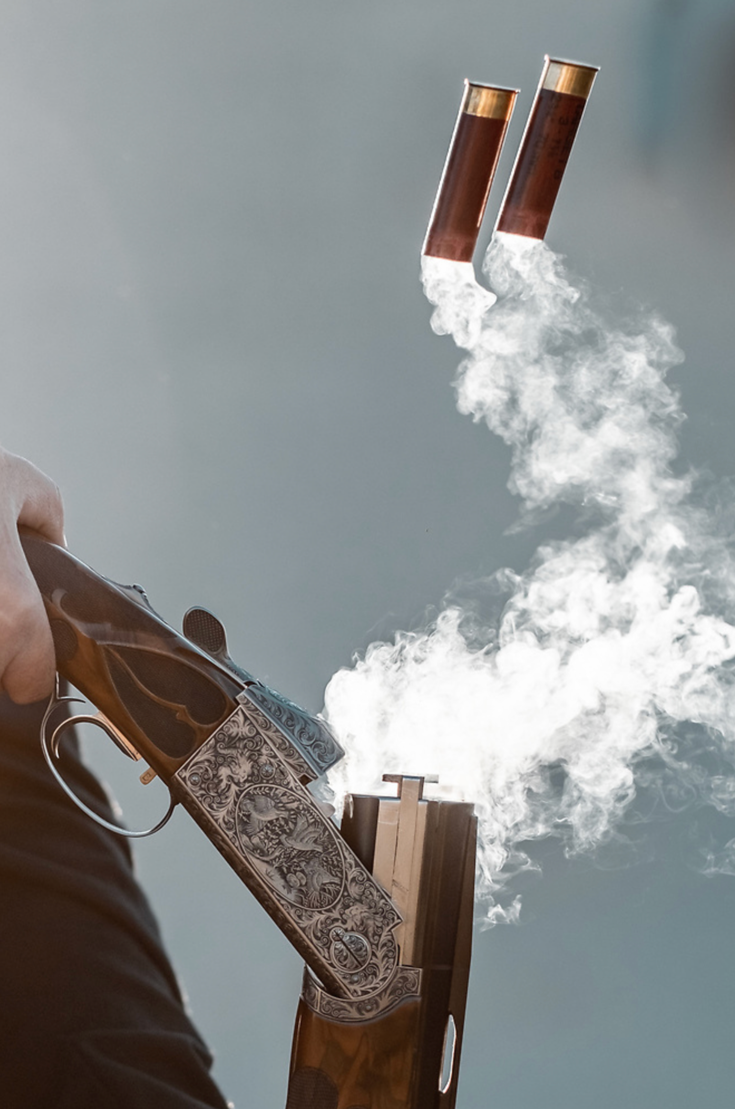 Smoke billows from a double-barrel shotgun against a grey sky background.