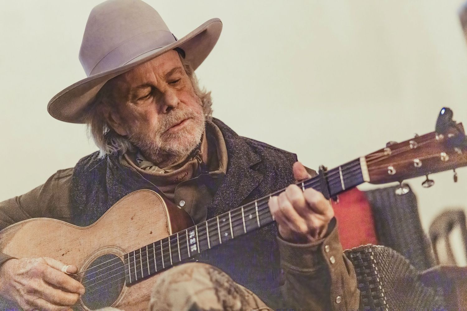 Musician playing acoustic guitar in warm lighting with cowboy hat against rustic backdrop.