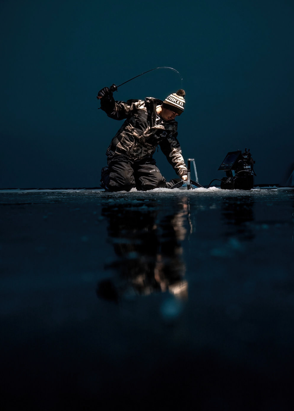 Ice fisherman in winter gear silhouetted against dark background while fishing on frozen lake.