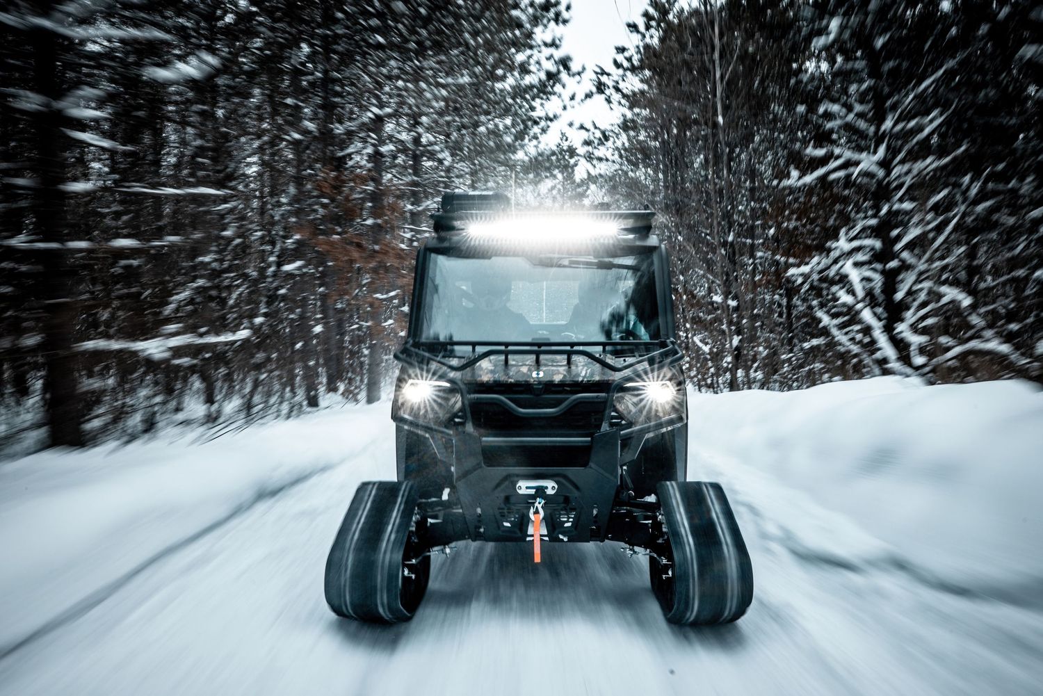 An off-road vehicle drives through a snowy forest path with bright headlights illuminating the way.