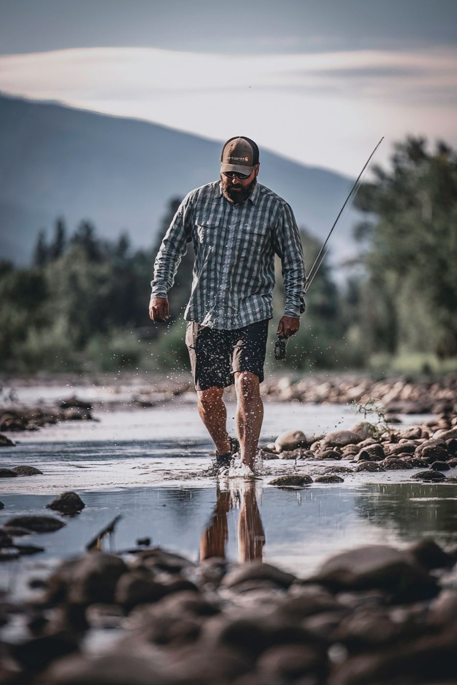 Angler standing in shallow rocky river water during sunset with fishing gear.