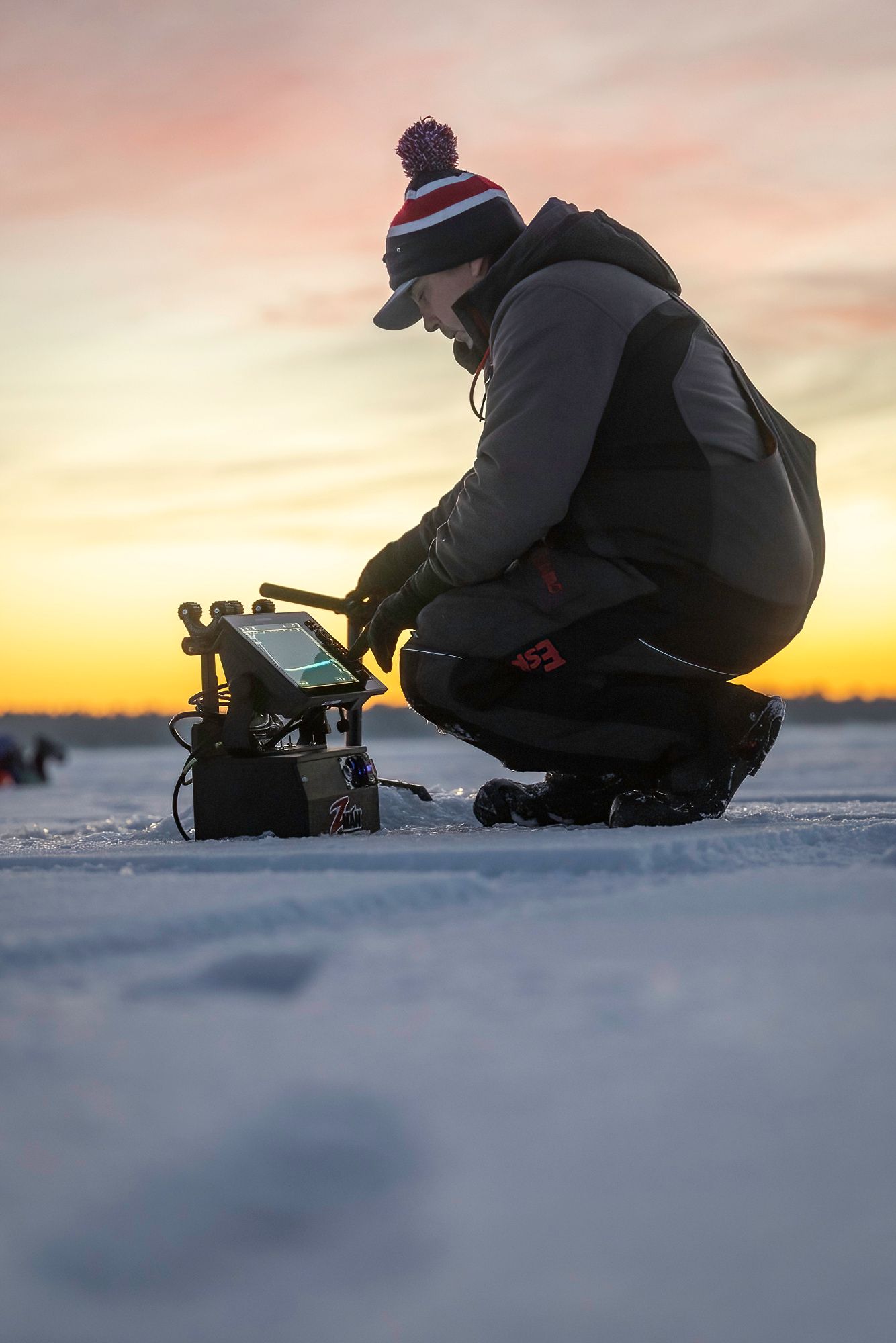 Ice fisher crouching to check equipment on frozen lake during colorful winter sunset.