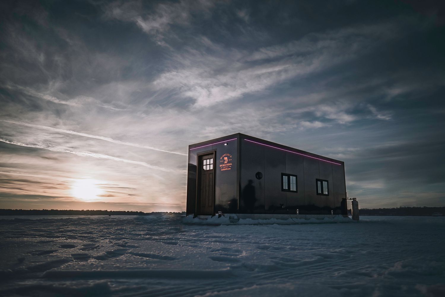 Ice fishing shelter on frozen lake with dramatic sunset and cloudy sky.