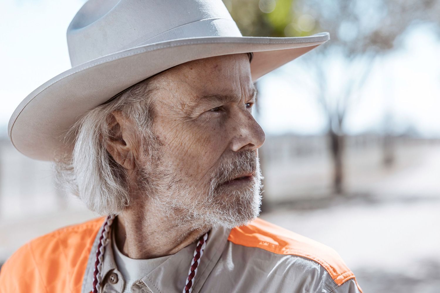Side profile of musician in cowboy hat and orange shirt performing outdoors.