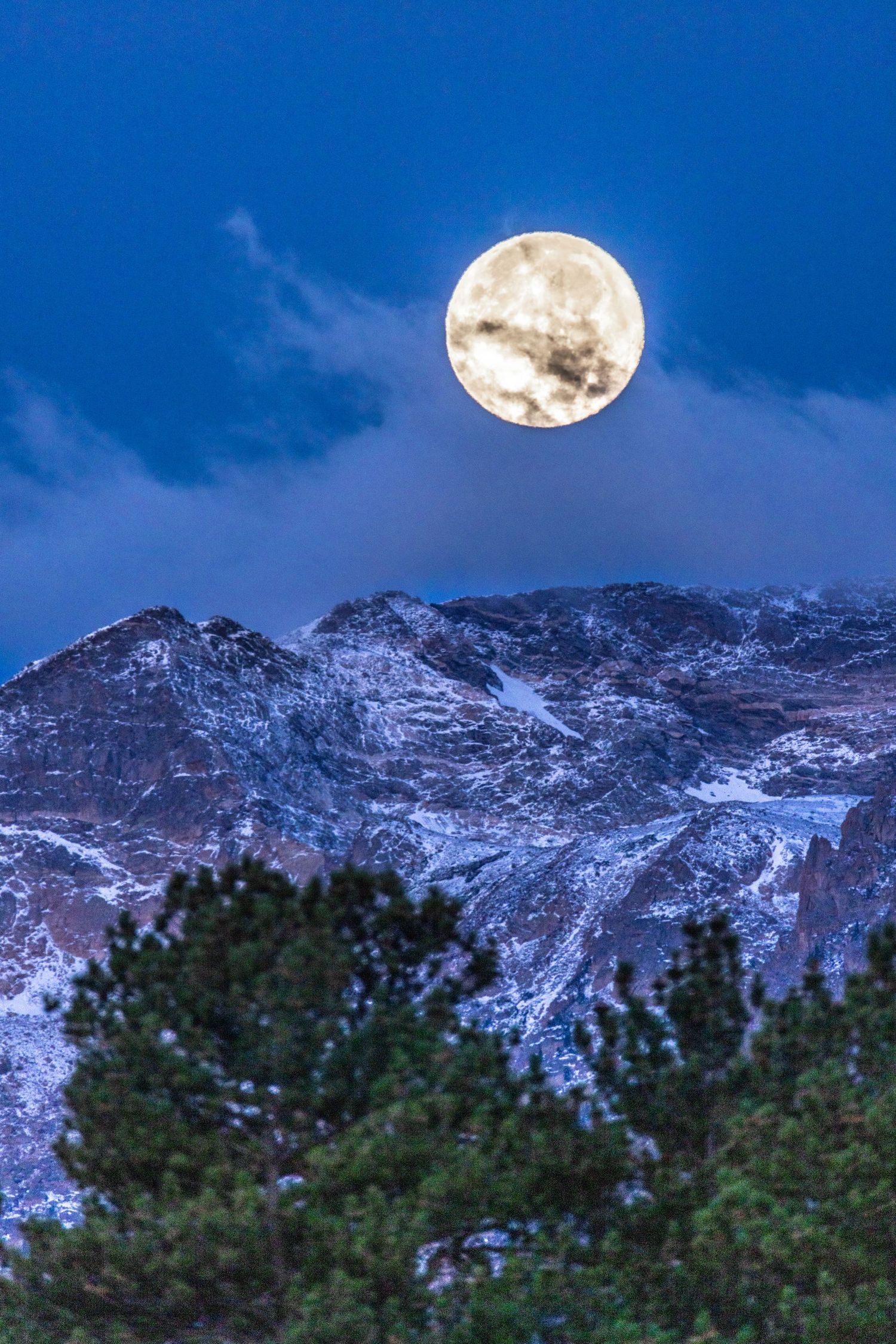 Full moon rises over snow-capped mountain peaks against deep blue evening sky.
