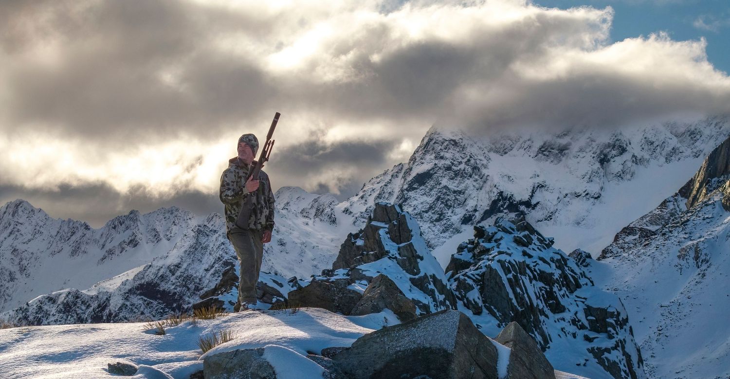 A hunter stands atop a snowy mountain peak against dramatic cloudy skies.