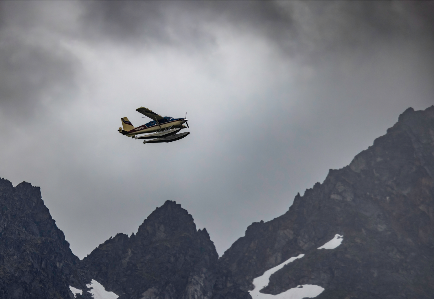 Small seaplane flying against dramatic mountain peaks in cloudy weather.