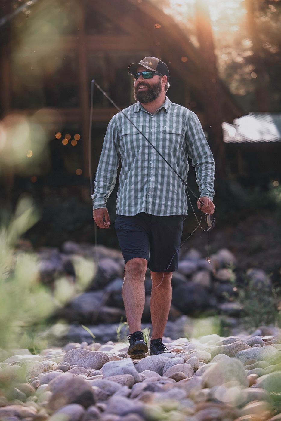 Fly fisherman walking on rocky riverbank during golden hour wearing plaid shirt and shorts.