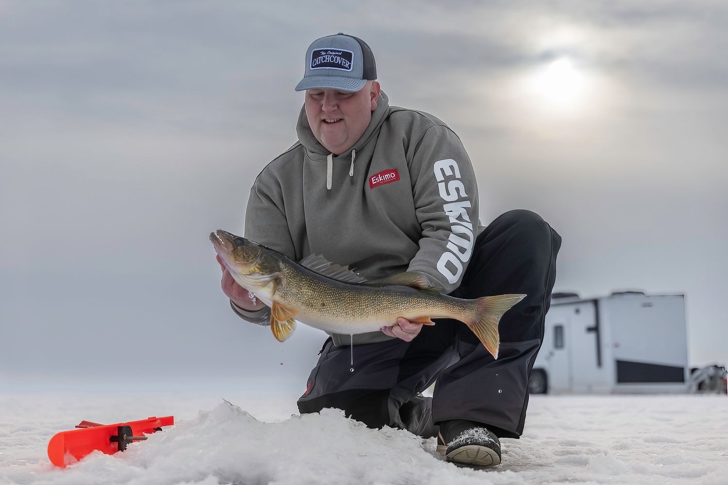 Ice fishermen set up on a frozen lake with fishing gear and ice houses visible in the overcast winter landscape.