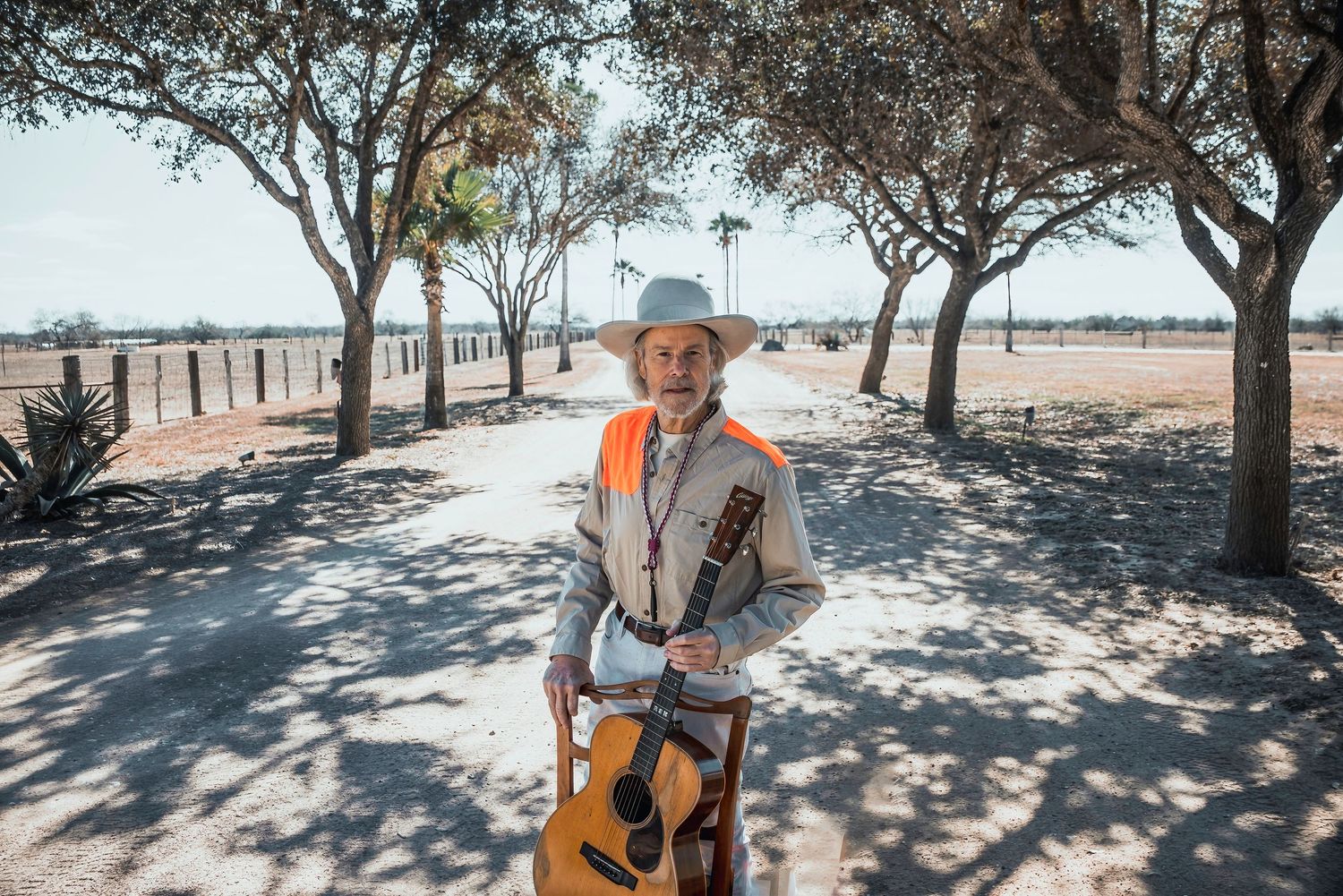 Guitarist performs in a rustic outdoor setting with trees casting dappled shadows.