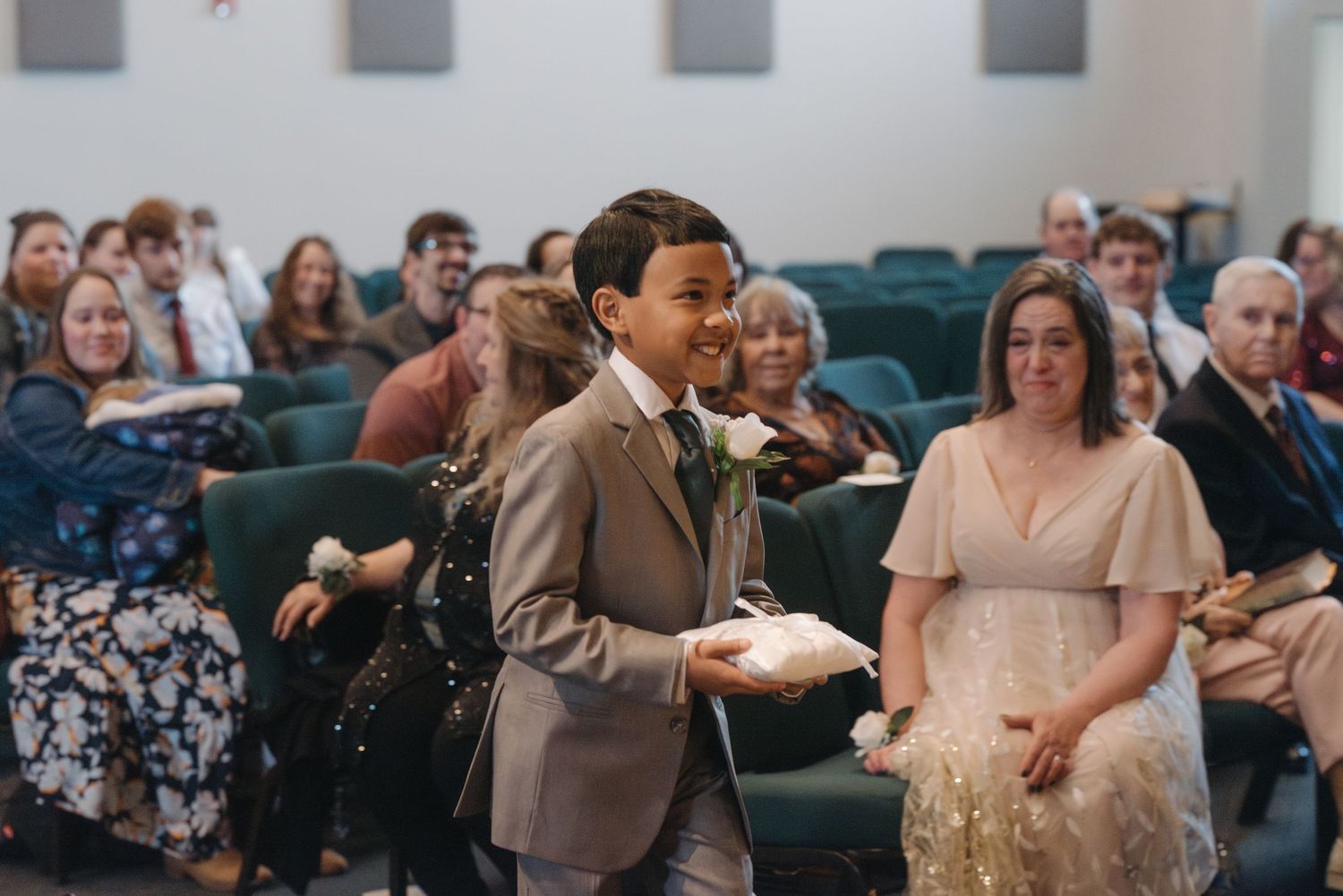 Smiling young boy in gray suit carries ring pillow down wedding aisle beside woman in champagne dress.