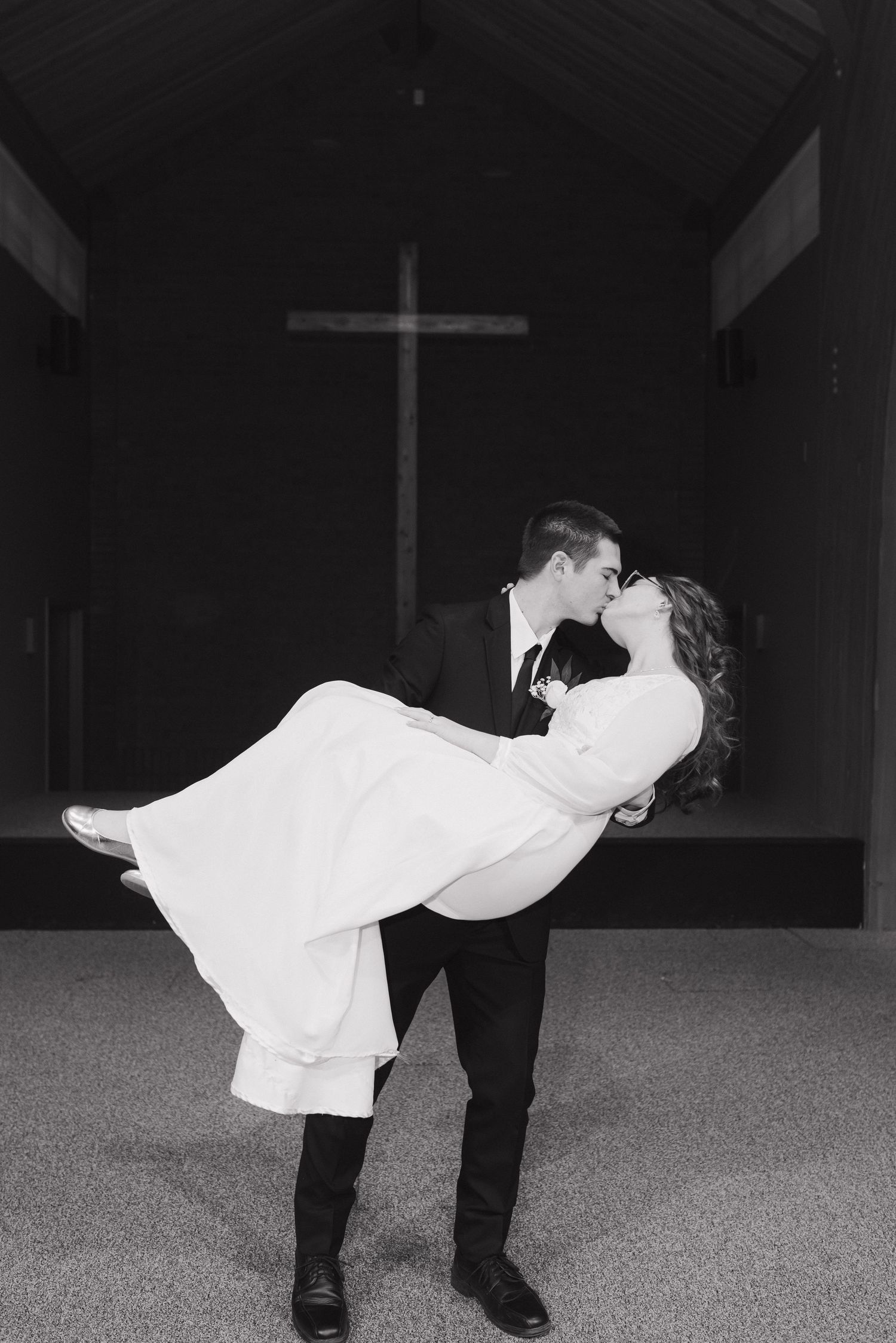 Groom lifts and kisses bride in white dress inside church chapel with large cross backdrop.