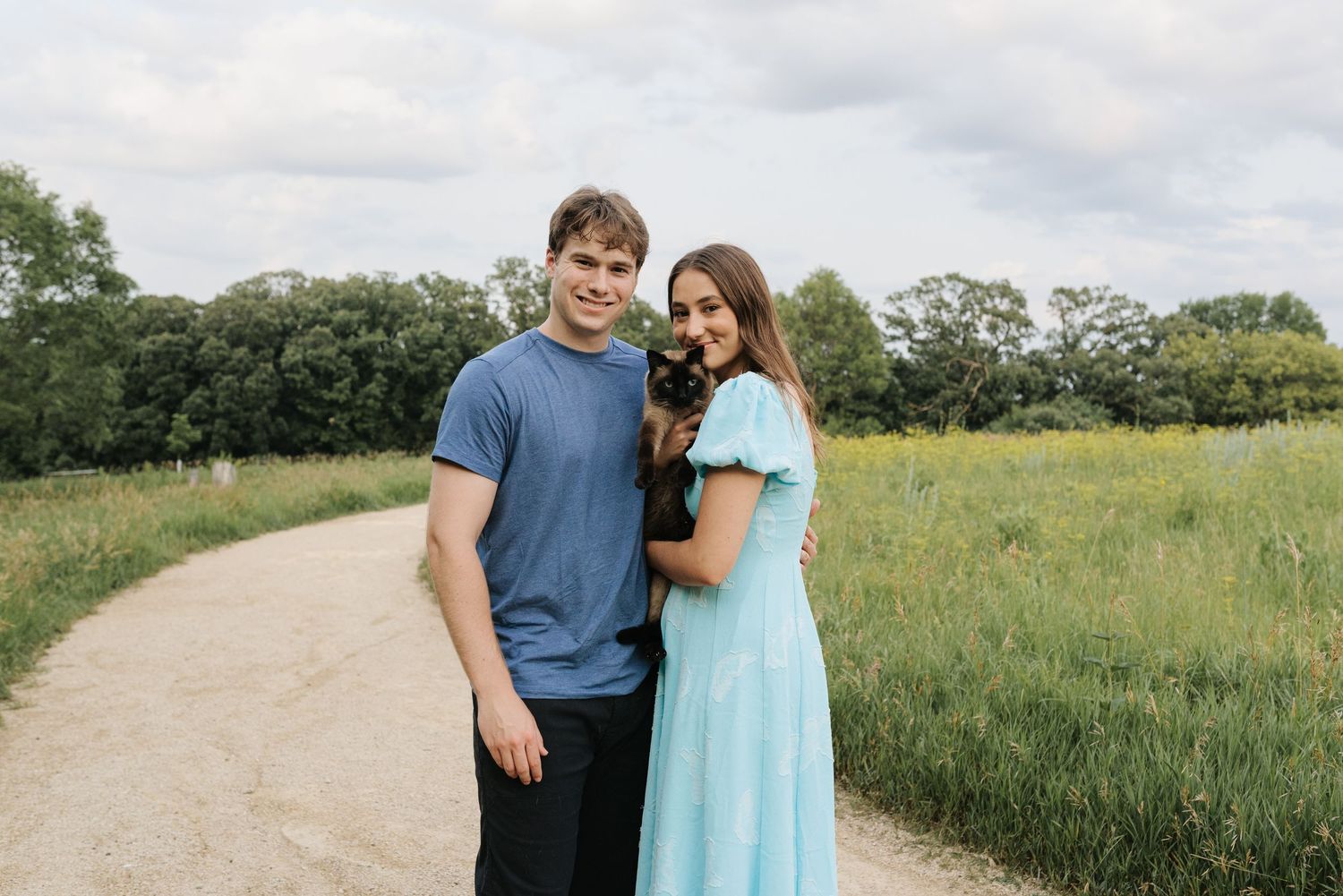 A smiling couple stands on a park path holding a small dog, surrounded by lush green fields under a cloudy sky.