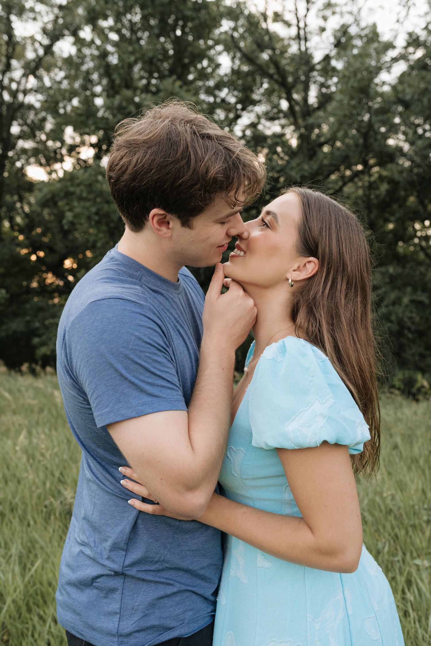 A young couple sharing a romantic moment outdoors, the man in a blue tee gently holding the woman in a light blue dress.