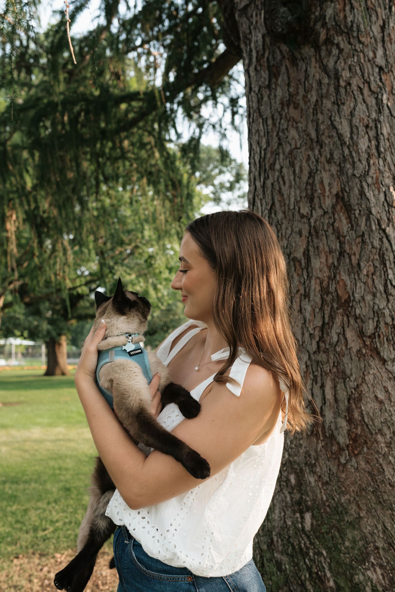 Woman in white top holds a Siamese cat outdoors near a large tree in a lush green park.