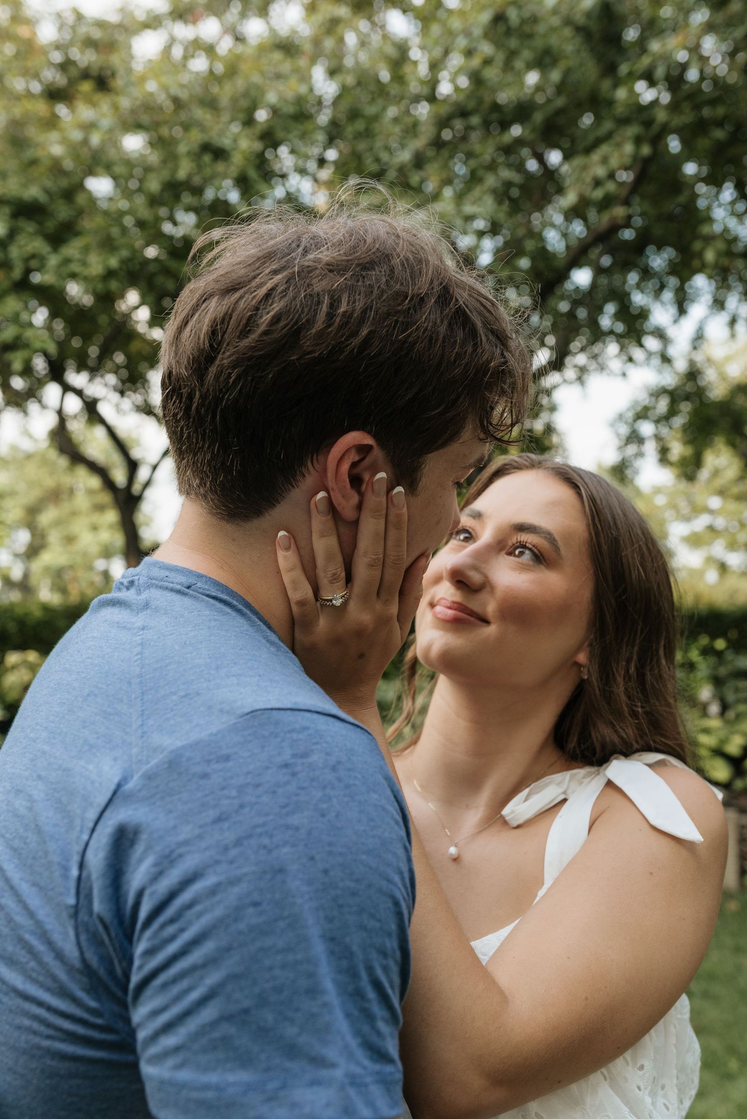 A smiling young woman gazes at a man in a blue shirt outdoors, with lush green trees in the background.
