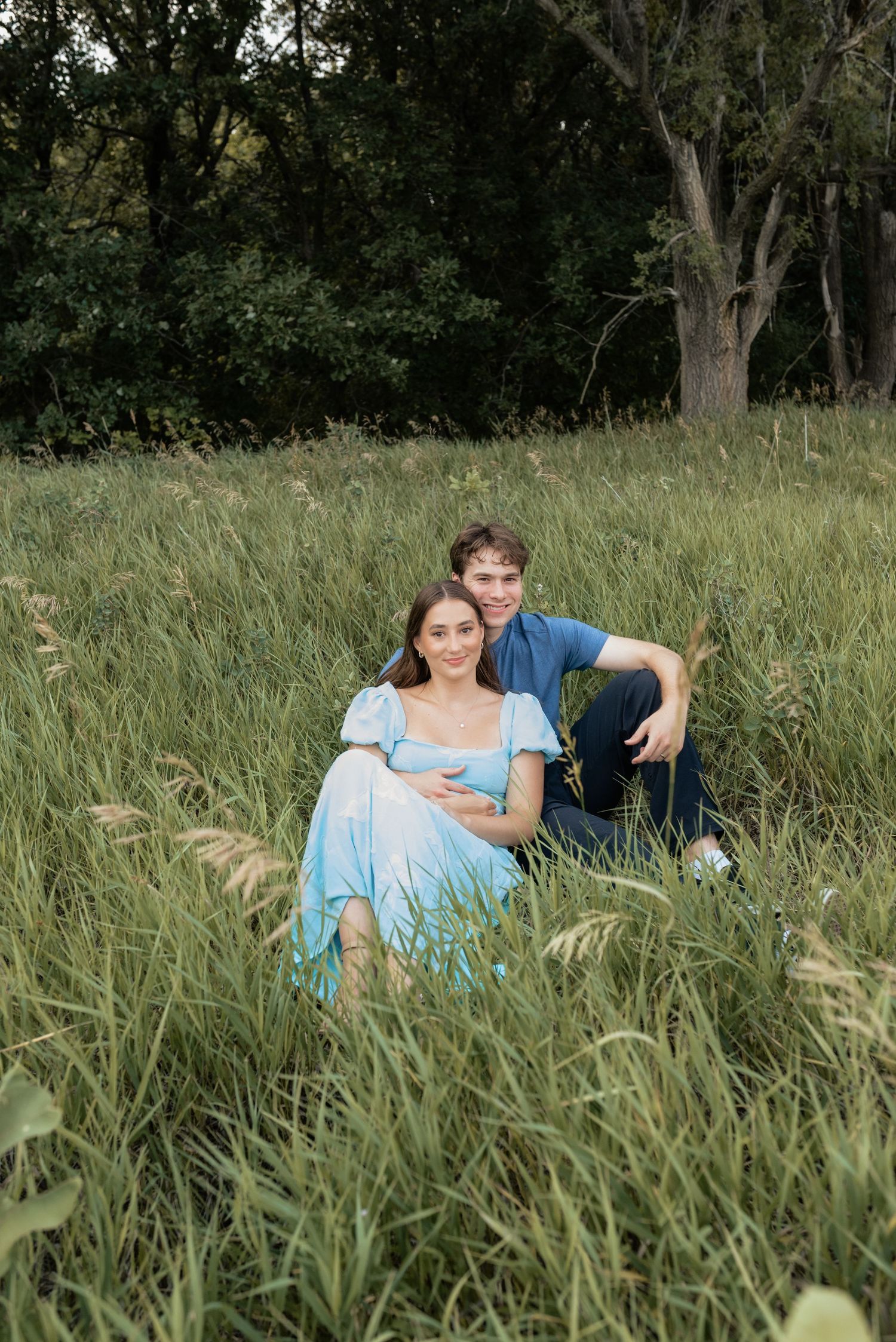 A couple sits together in tall green grass, the woman in a blue dress cradling her baby bump, surrounded by lush trees.