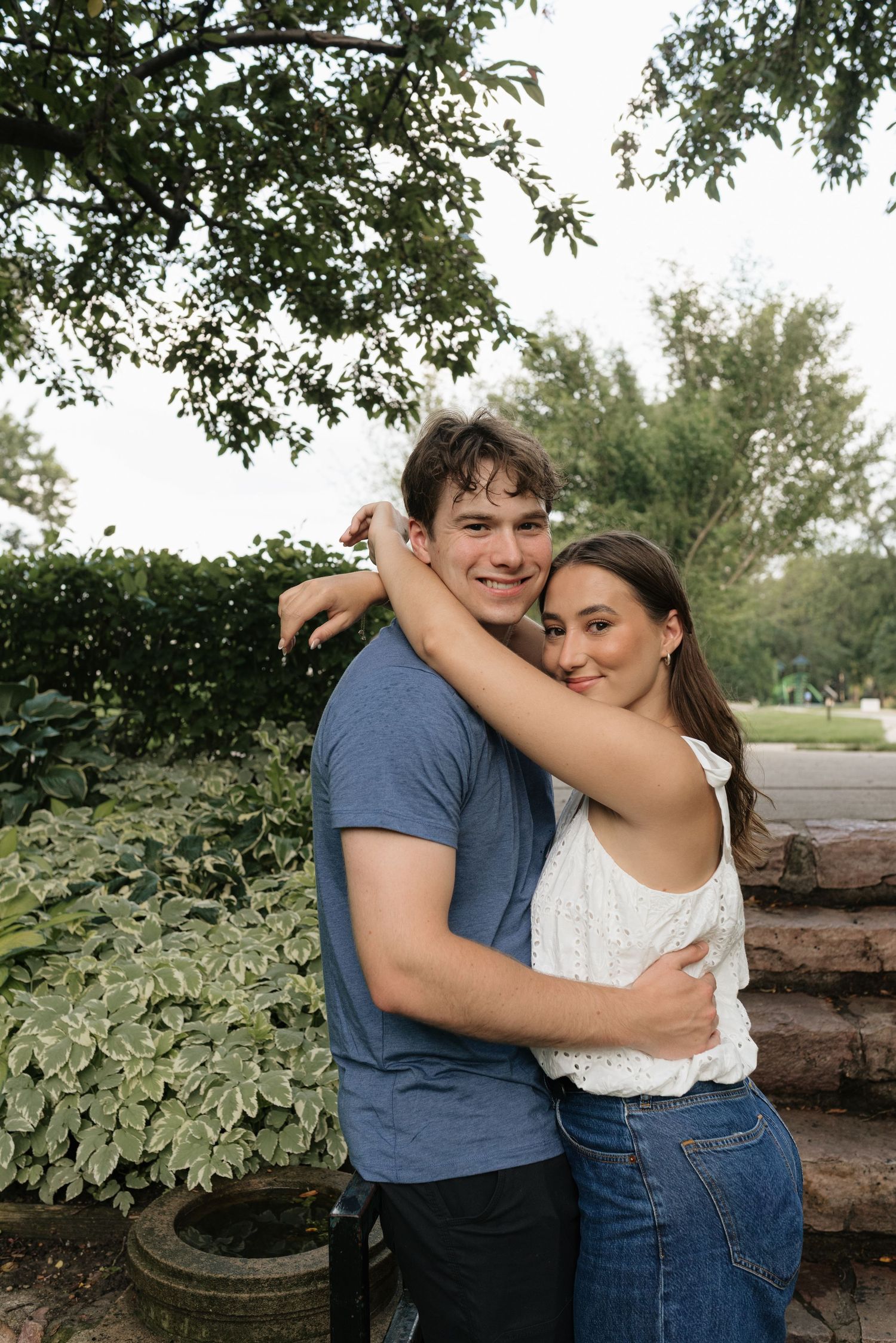 A young couple embracing and smiling outdoors, surrounded by lush green plants and trees.