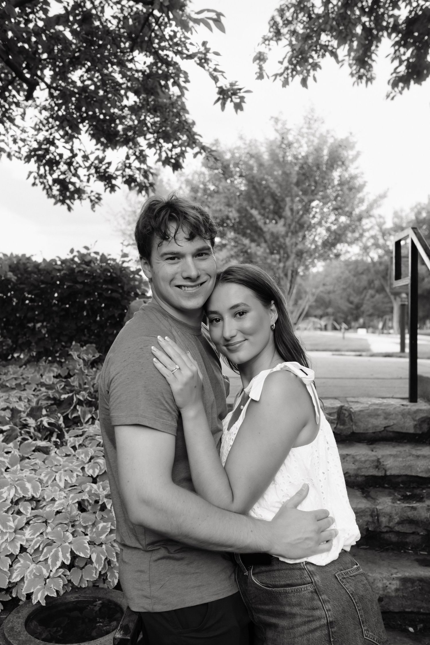 Black and white photo of a smiling couple embracing outdoors near lush trees and garden steps, woman showing engagement ring.