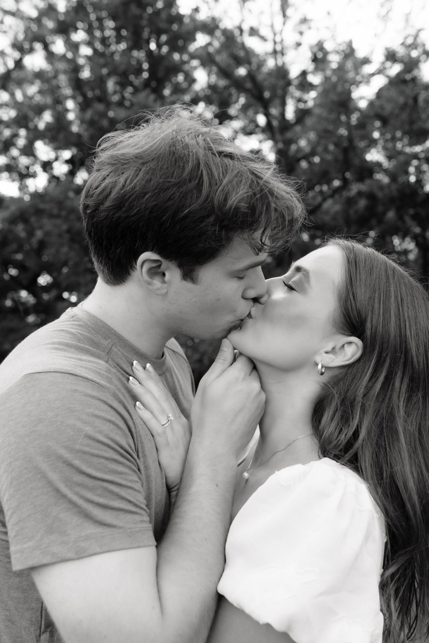 Black and white photo of a romantic couple sharing a tender kiss outdoors with blurred trees in the background.