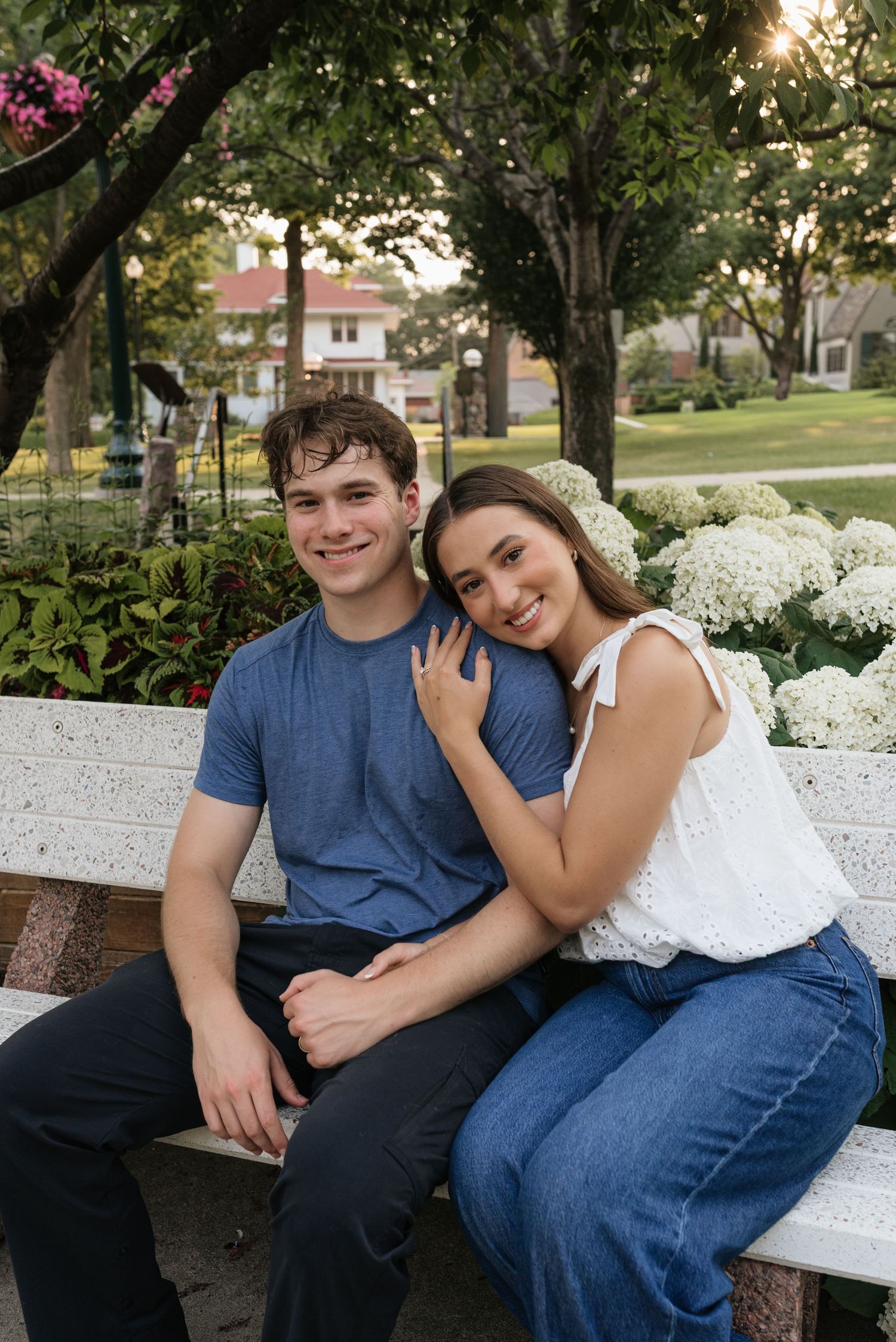 Young couple sitting on a white bench in a park, smiling, surrounded by white hydrangeas and green trees.