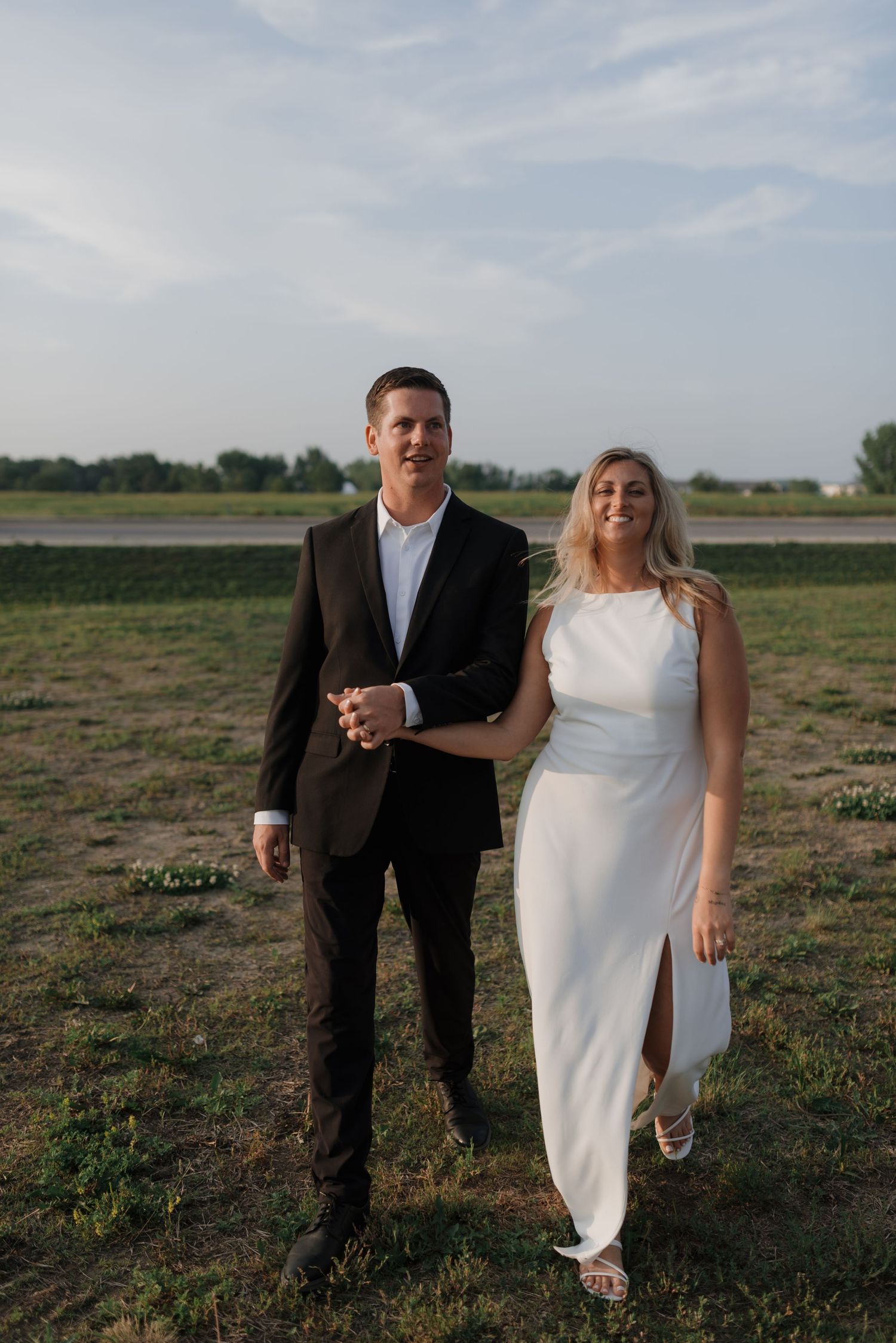 Cheerful bride and groom smiling and walking together across a green field at sunset.