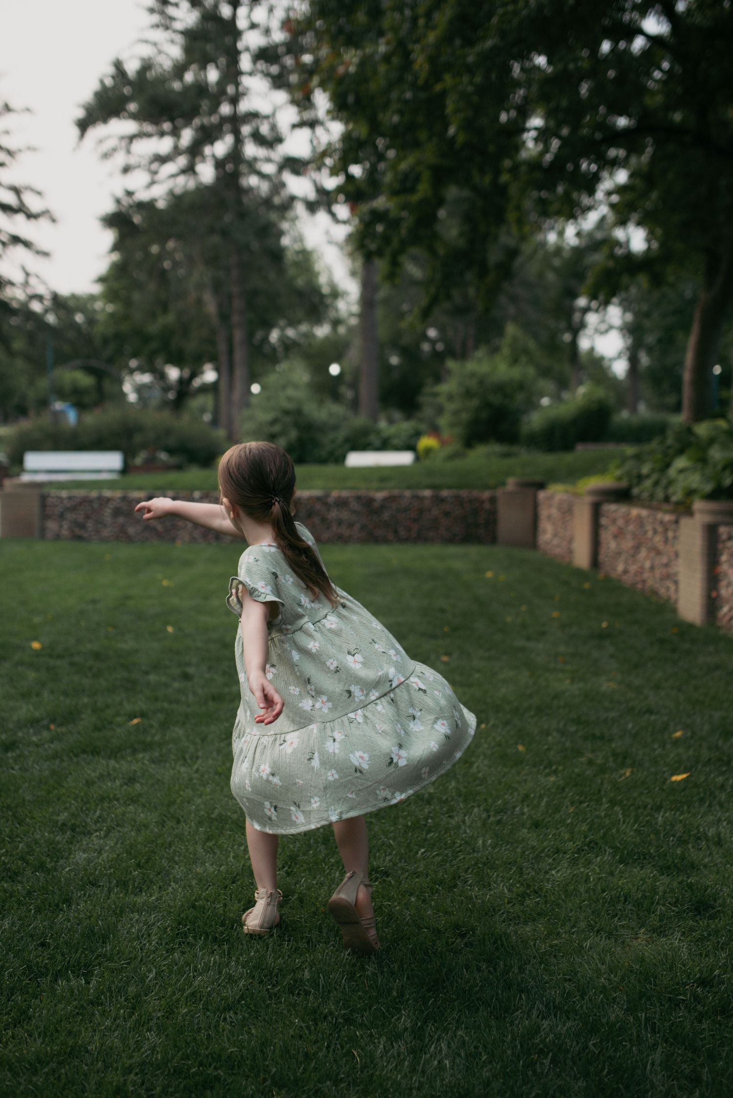 Little girl in light green floral dress spinning outdoors on green grass in a park.