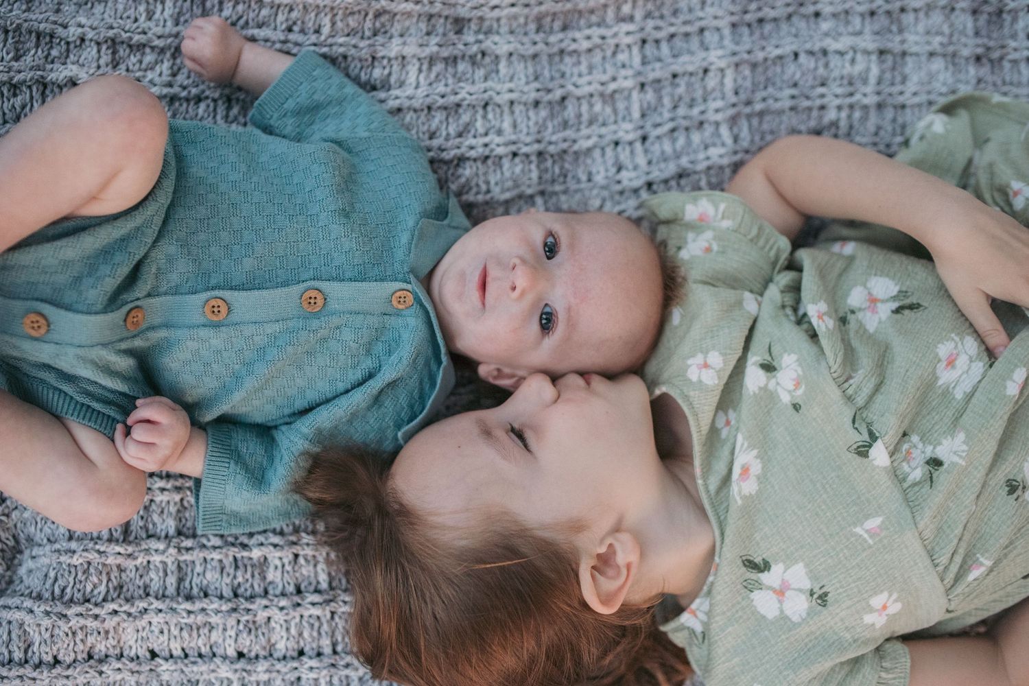 Sweet baby siblings lying together on gray blanket, older girl in floral dress smiling.