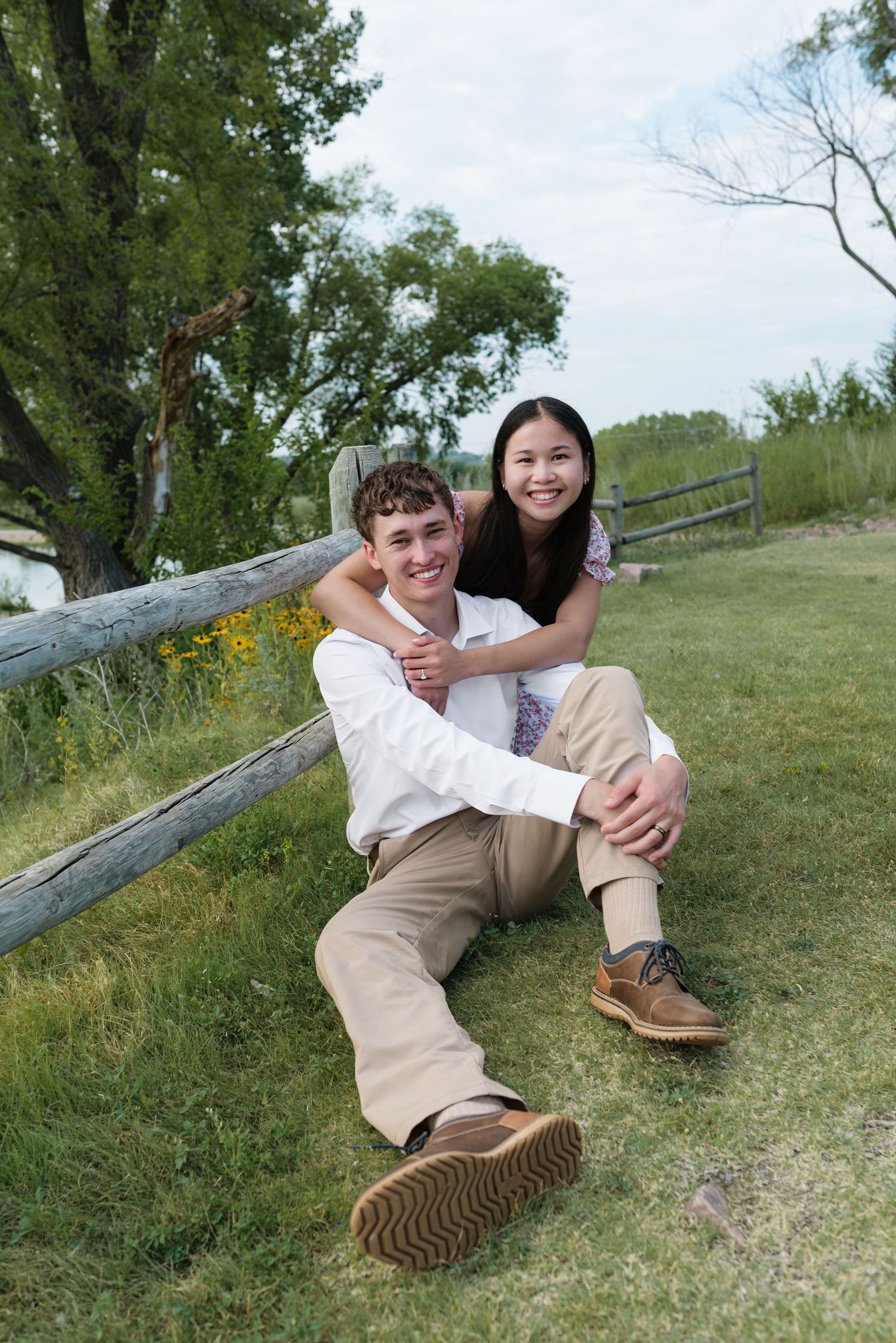 Happy couple sitting on grass by a wooden fence, smiling outdoors in a lush green park.