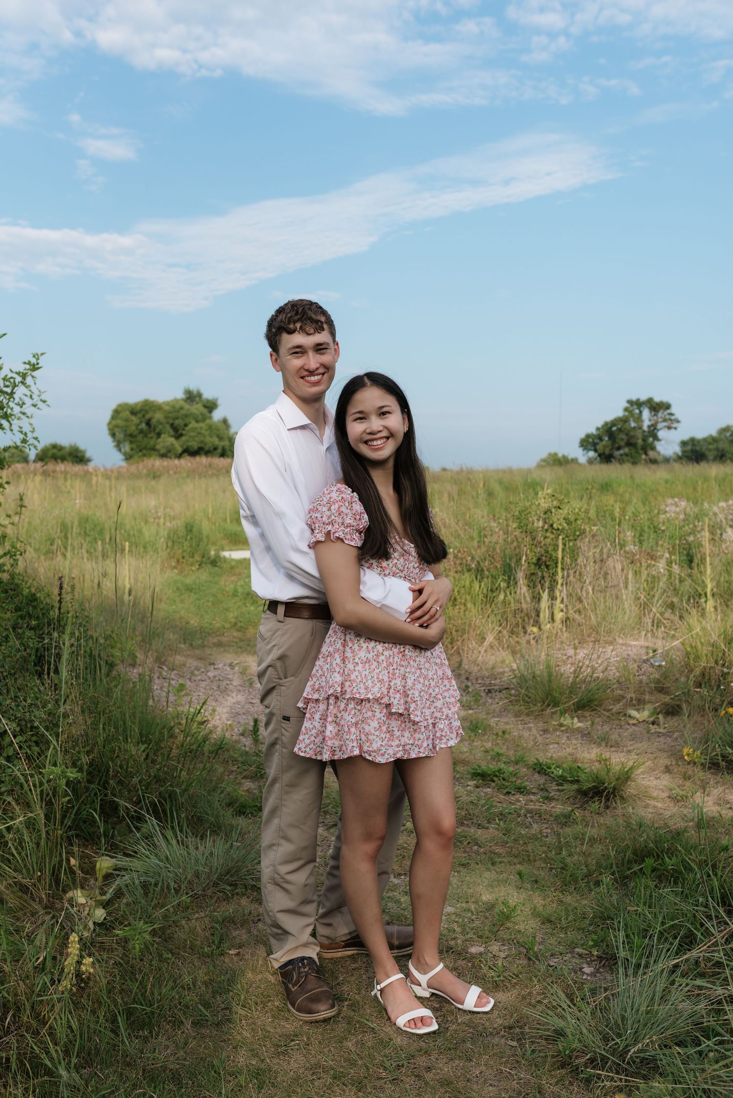 A smiling couple embraces on a grassy outdoor path under a bright blue sky in a natural meadow.
