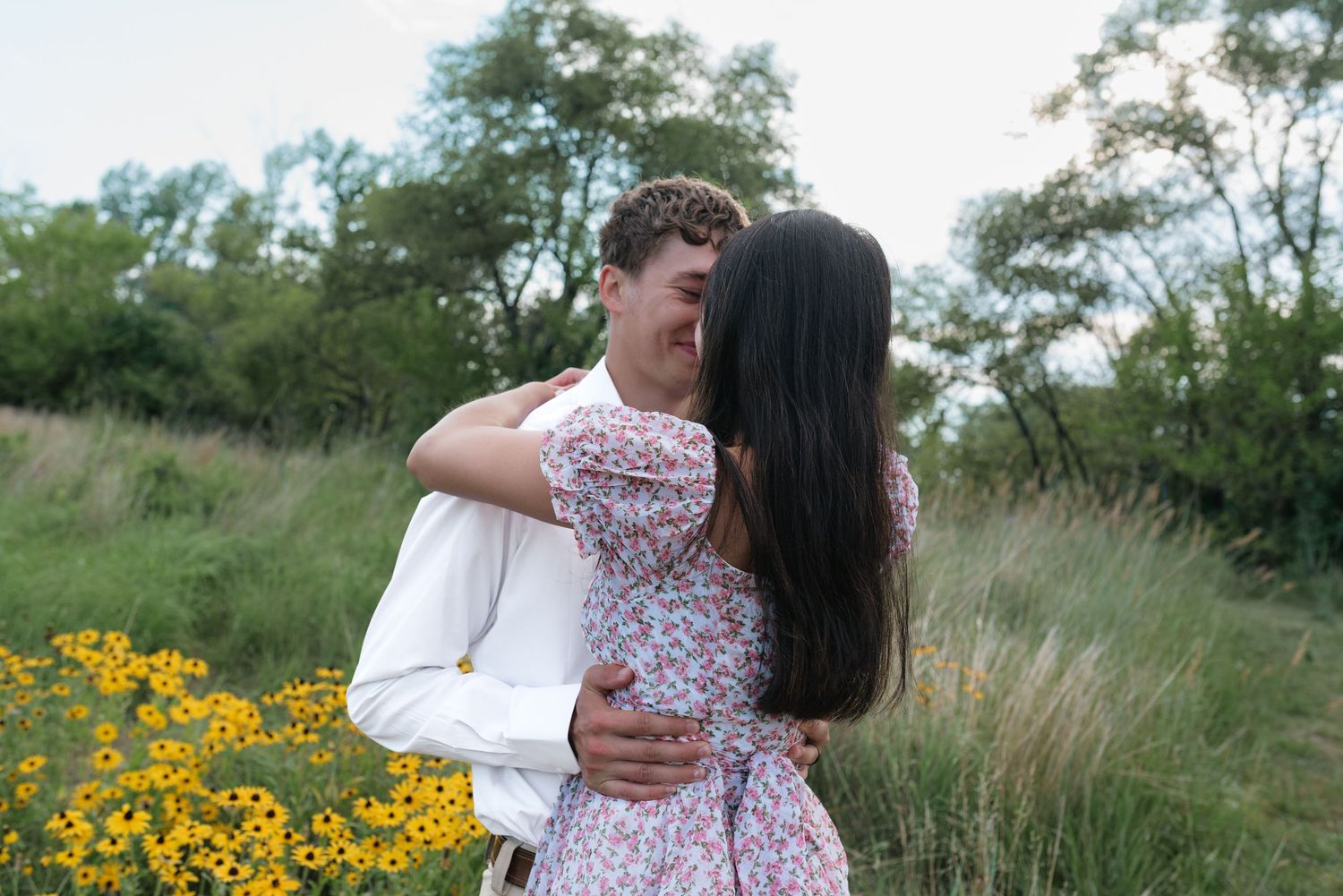 A couple embraces and kisses in a lush green field with yellow wildflowers, woman in floral dress, man in white shirt.