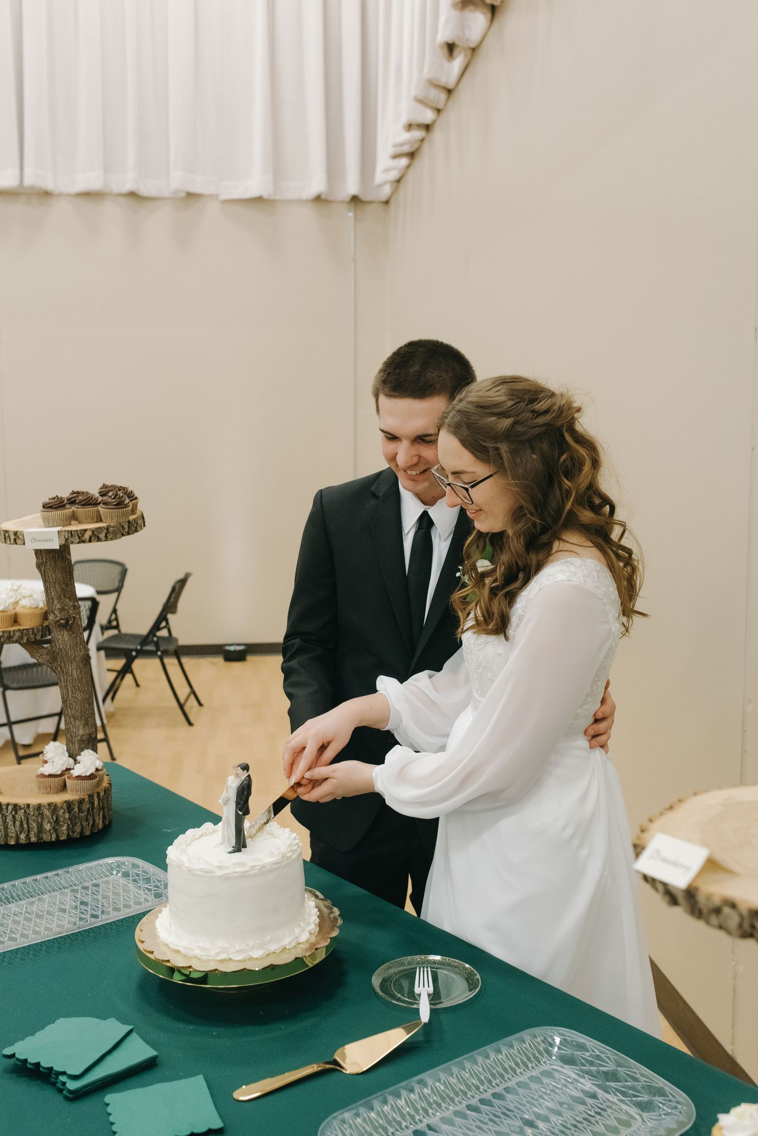 Newlyweds smiling while cutting white single-tier wedding cake at green-draped reception table.