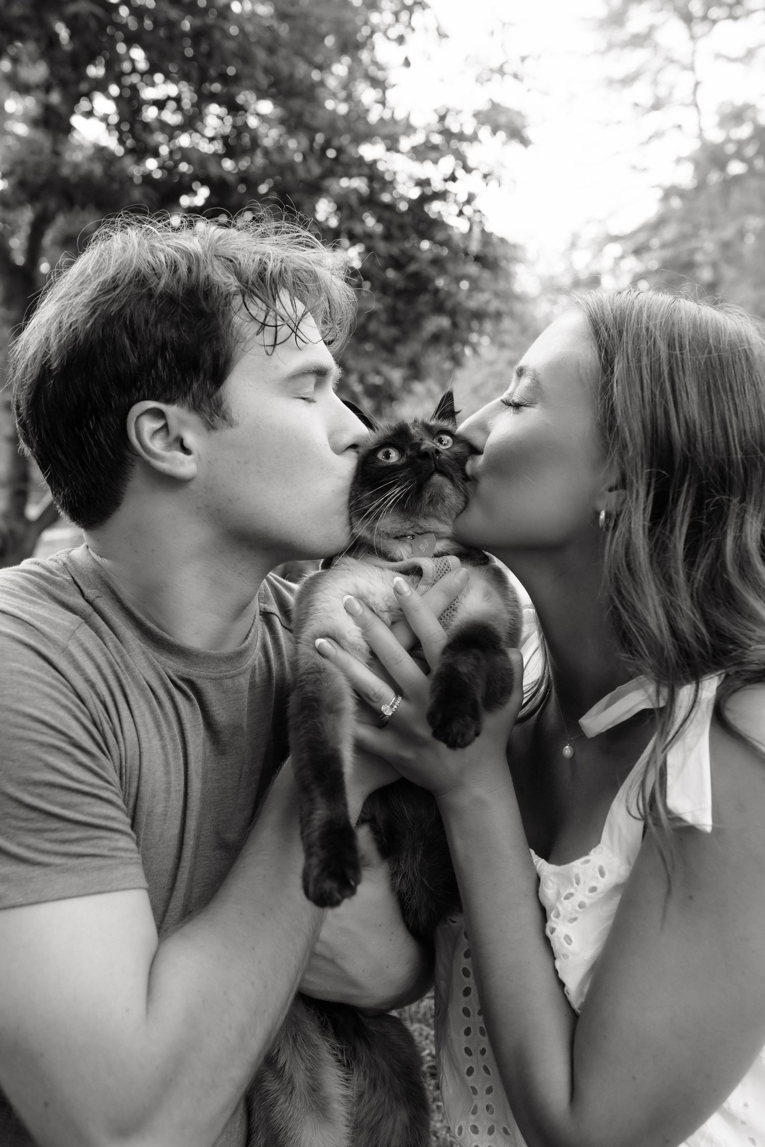 Black and white photo of a couple kissing their Siamese cat outdoors, surrounded by trees.