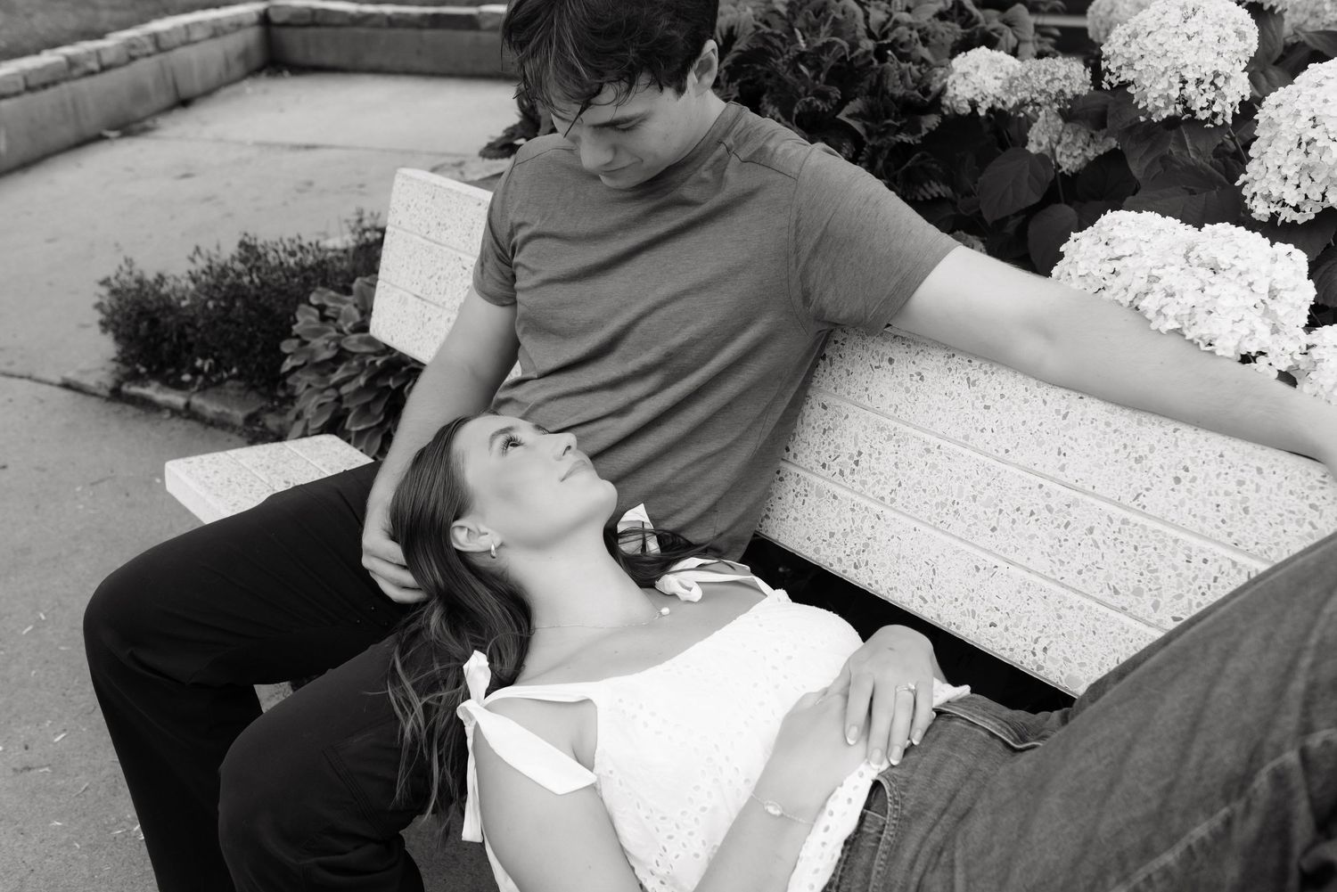 Black and white photo of a romantic couple relaxing together on a stone bench surrounded by blooming hydrangeas.